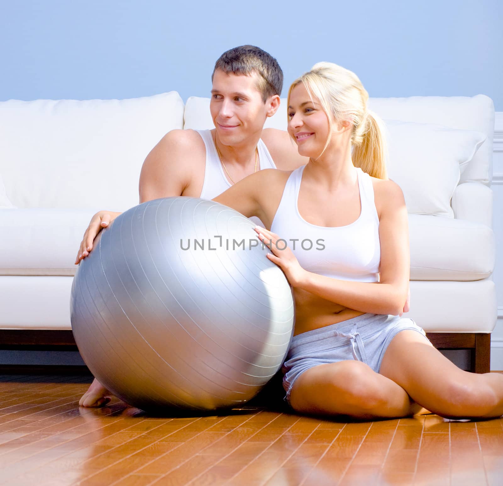 Young couple sit on the wood floor, smiling off to the side while holding a silver exercise ball. Horizontal shot.