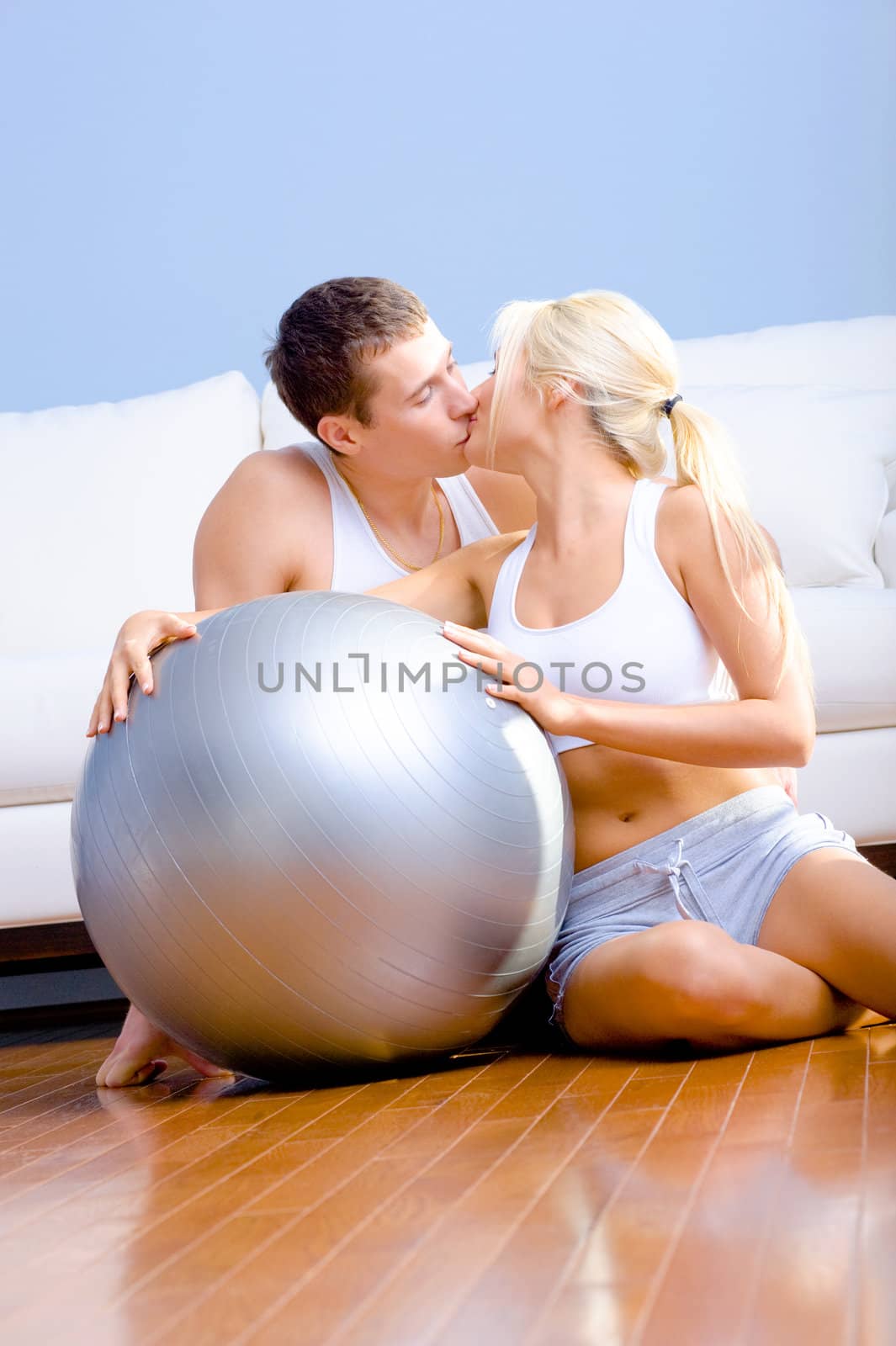 Young couple in sportswear kiss while sitting next to an exercise ball. Vertical shot.