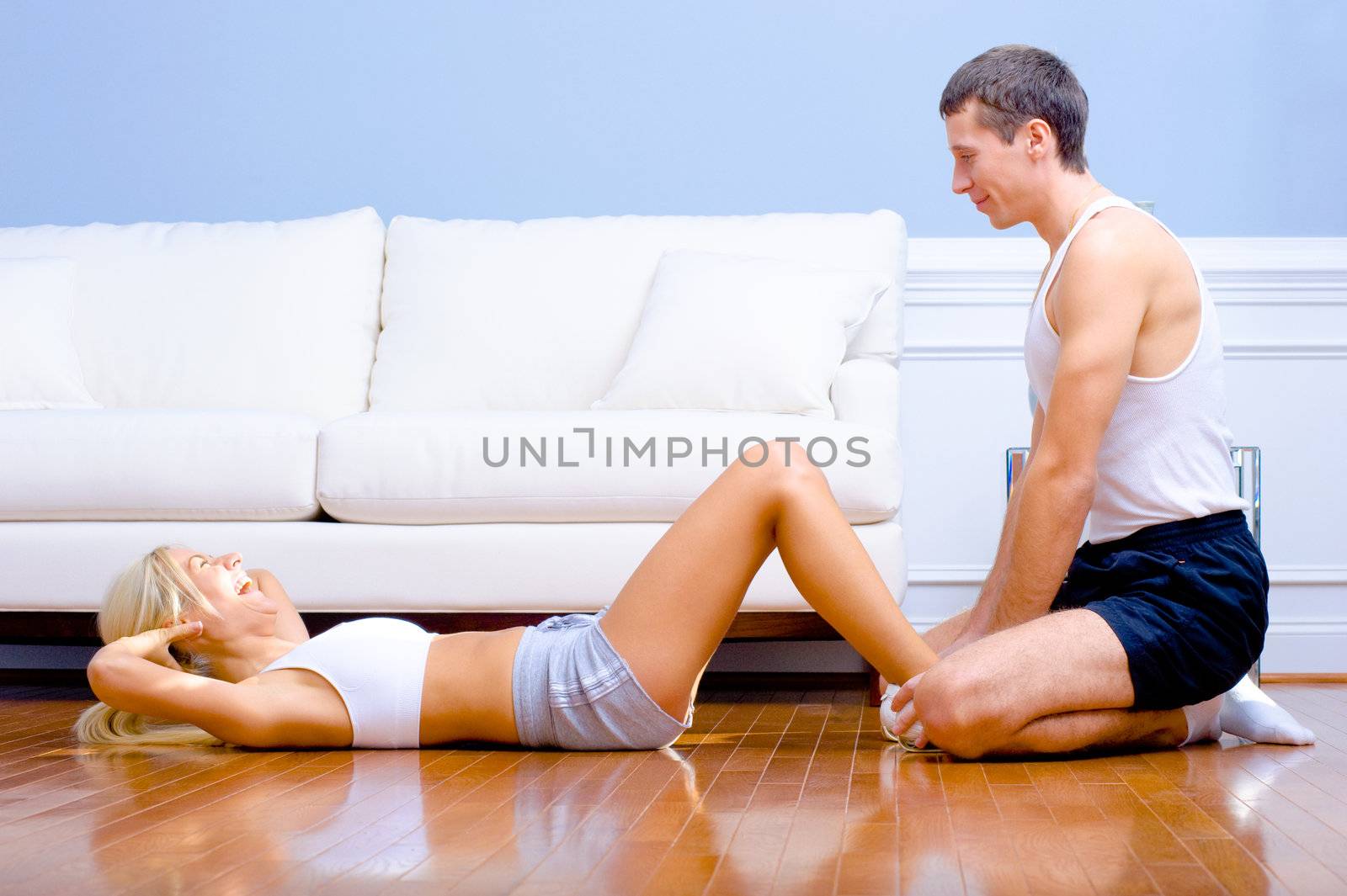 Young woman laughs while doing sit ups on the wood floor.  A young man is holding her feet. Horizontal shot.