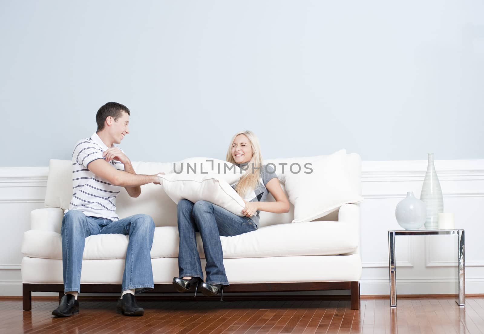 Young man playfully hits a young woman with a pillow on the sofa. Horizontal shot.