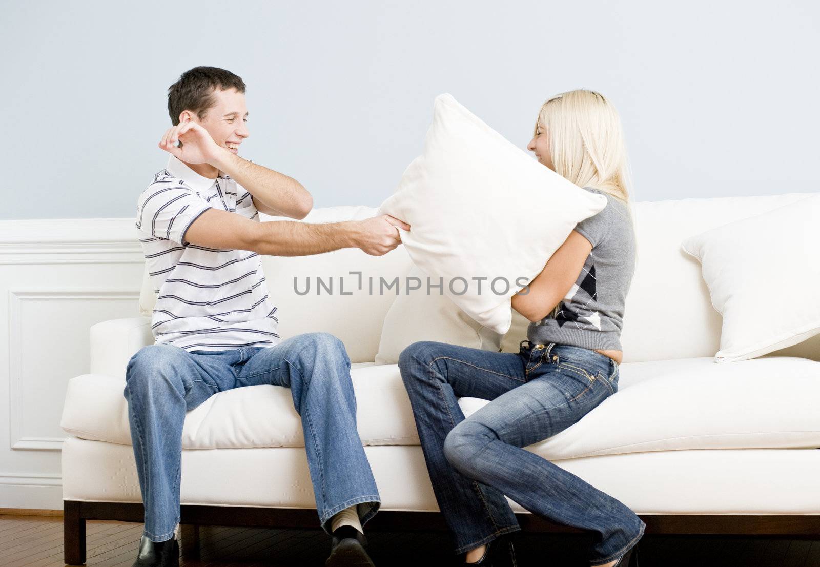 Young couple laugh while having a pillow fight on the sofa. Horizontal shot.