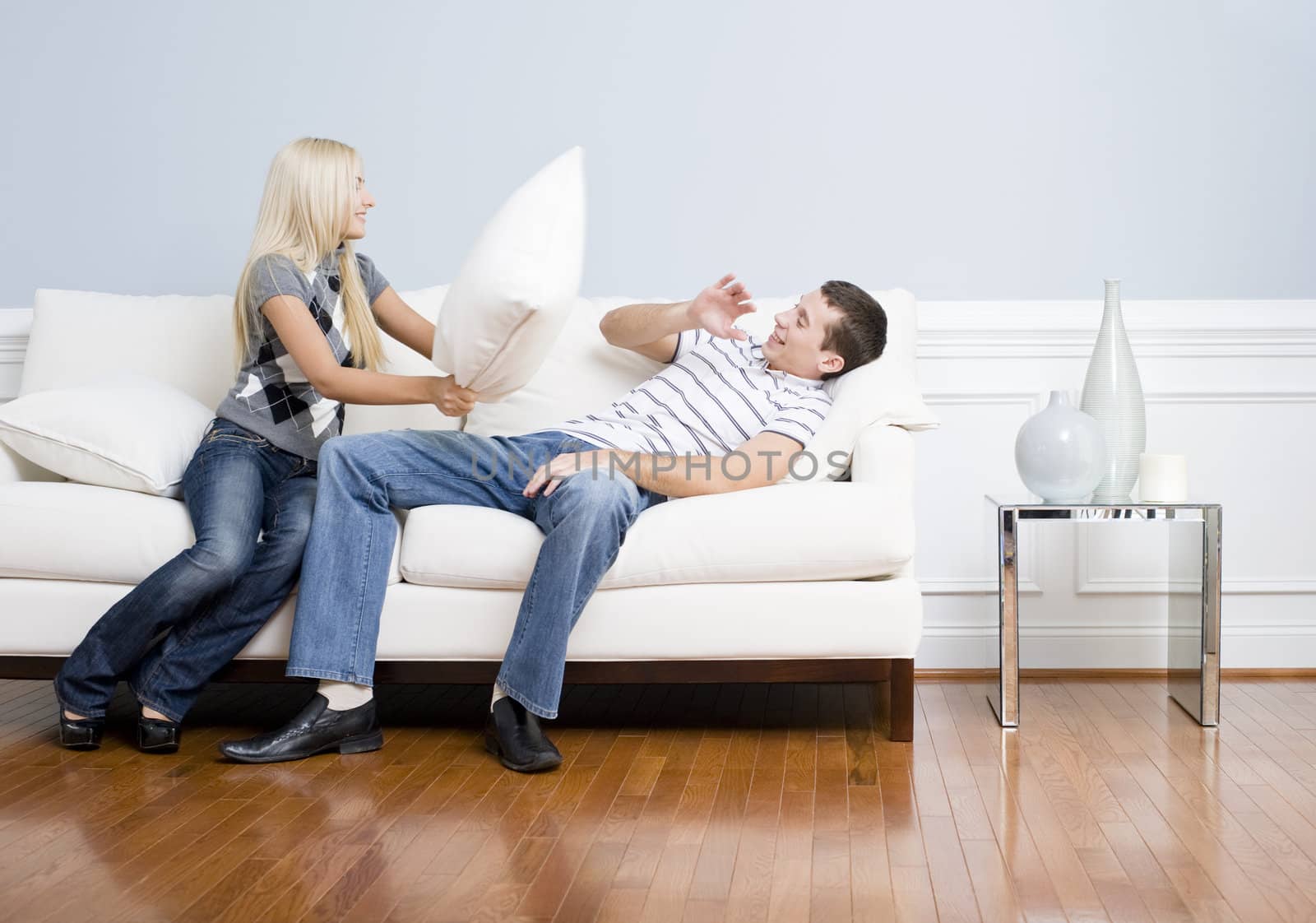 Young couple playfully have a pillow fight on a sofa.  The young man tries to avoid getting hit. Horizontal shot.