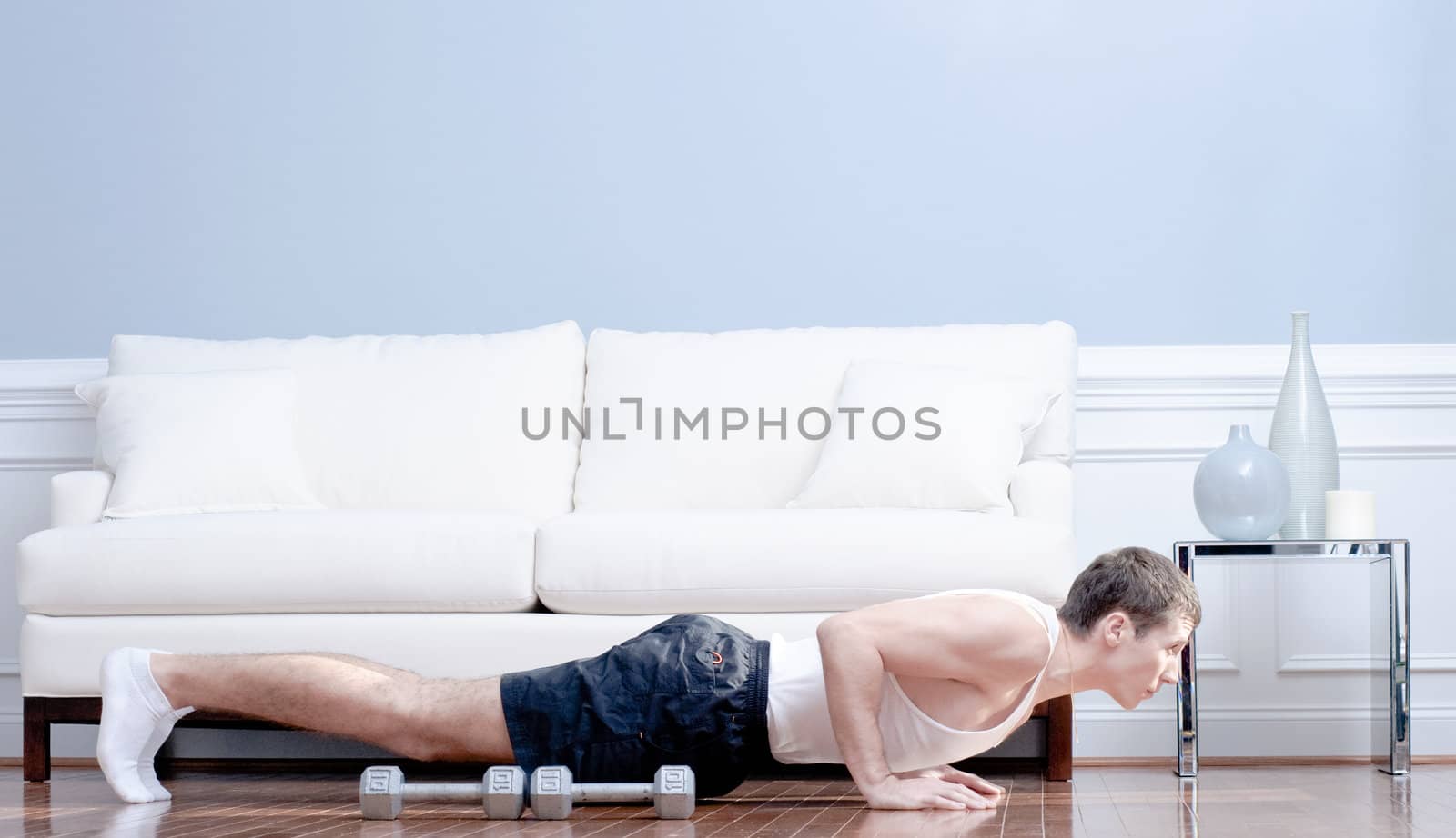 Full length view of man doing push-ups next to couch, with arm weights lying next to him. Horizontal format.