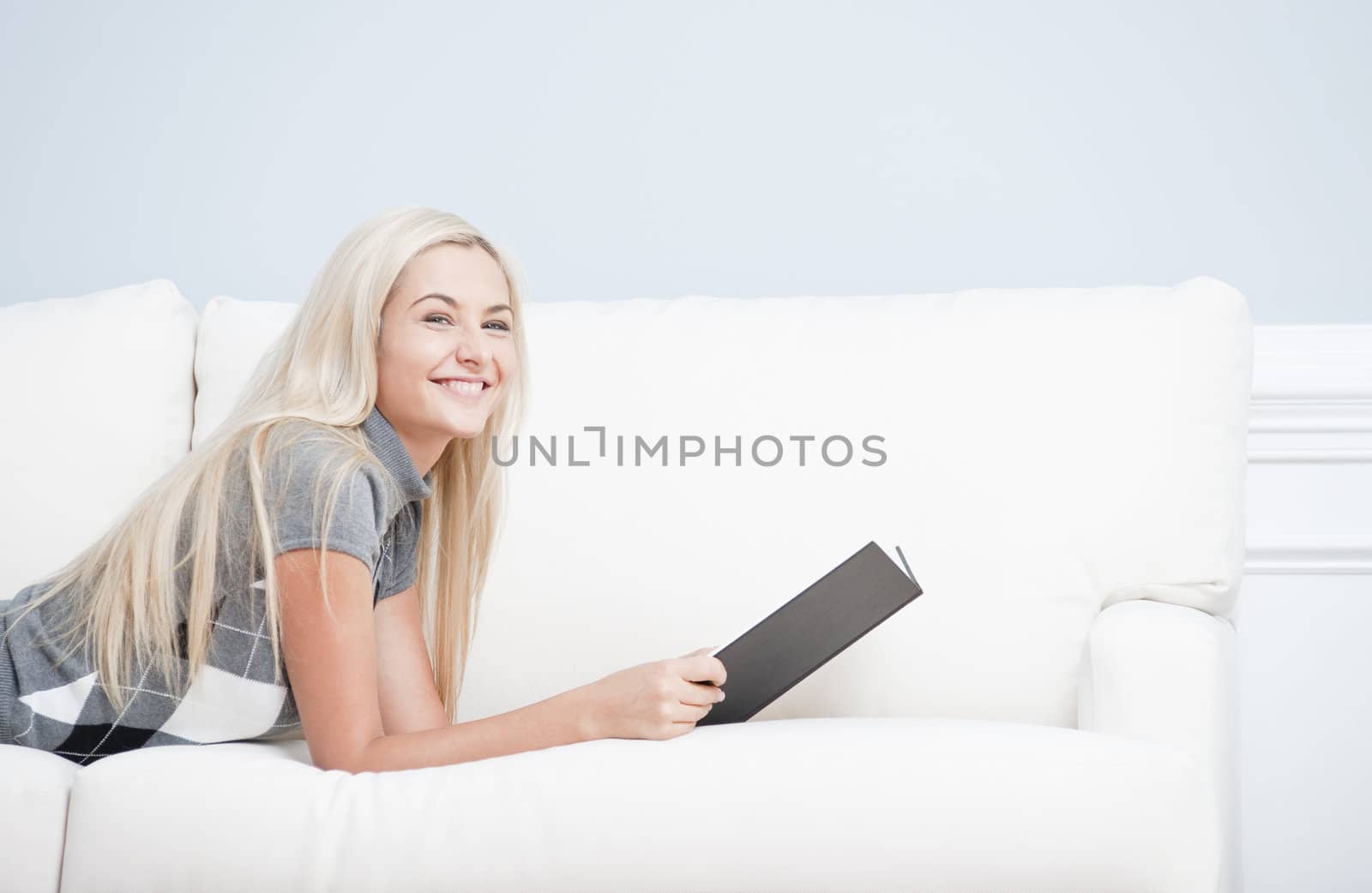 Cropped view of woman reclining on white couch with book and looking up with a smile. Horizontal format.