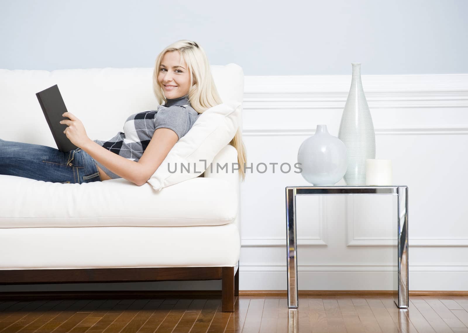 Cropped view of a woman relaxing on a white couch with a book and smiling at the camera. Horizontal format.