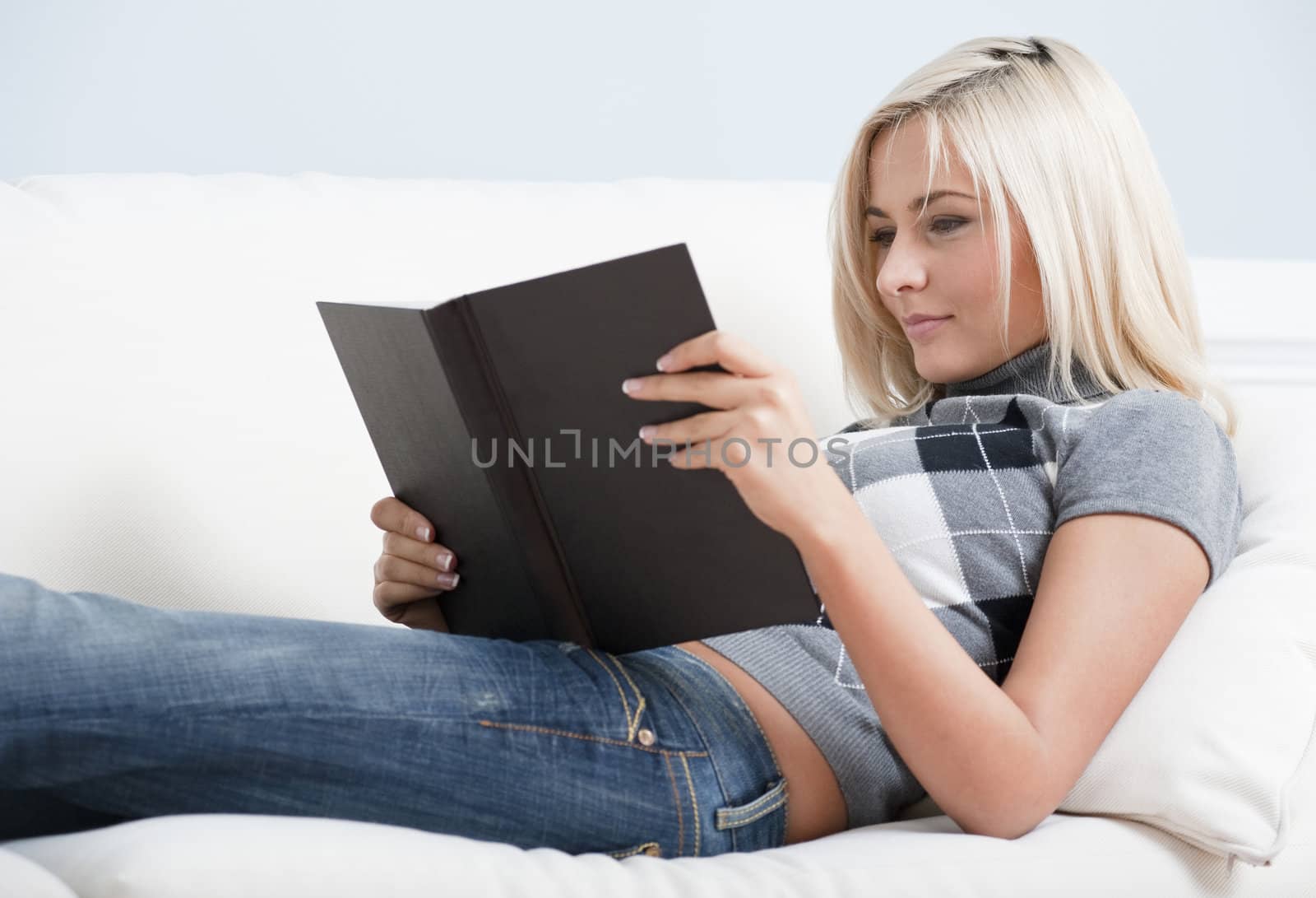 Cropped view of smiling woman relaxing on white couch with a book. Horizontal format.