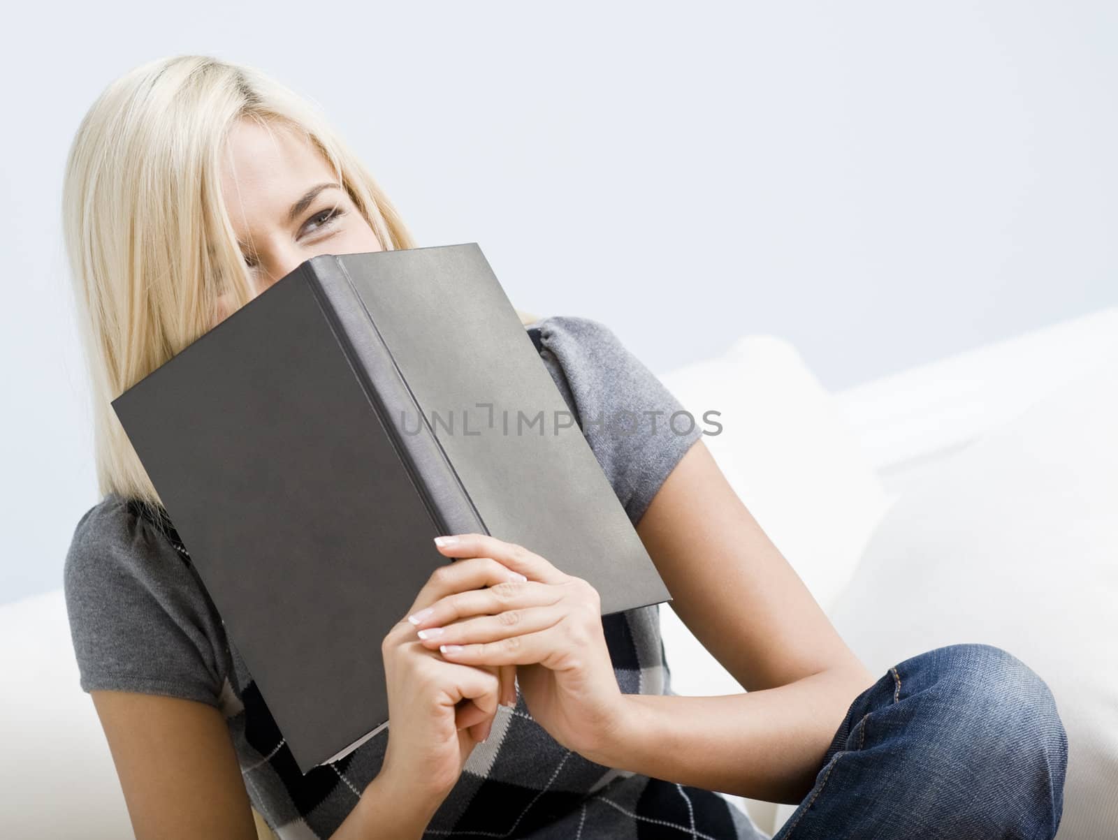 Tilt view close-up of woman sitting on white couch and giggling as she hold a book up to her face. Horizontal format.