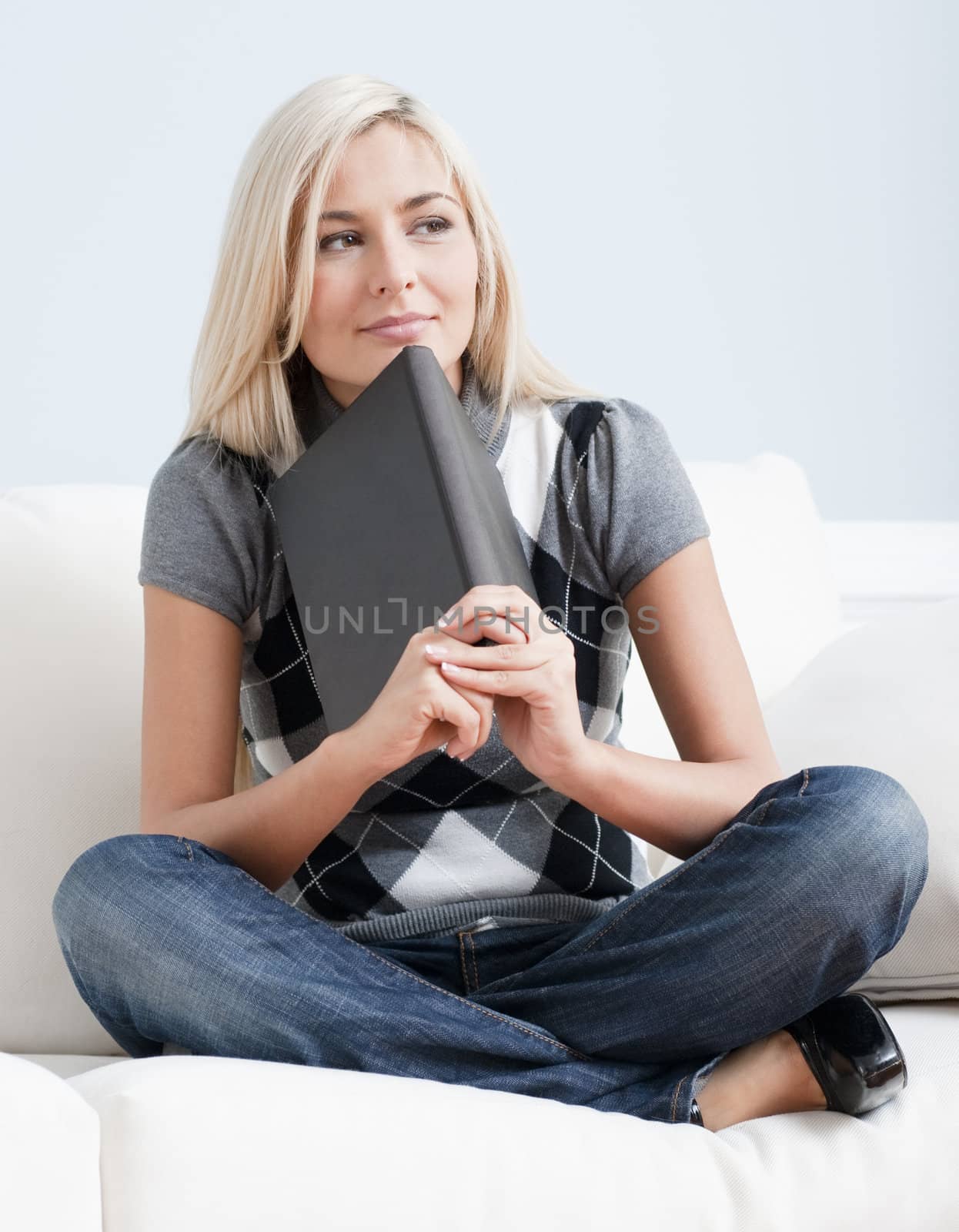 Contemplative woman sitting cross-legged on white couch and resting her chin on a book. Vertical format.