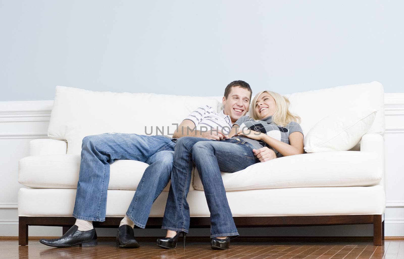 Full length view of affectionate couple laughing and relaxing together on white couch. Horizontal format.