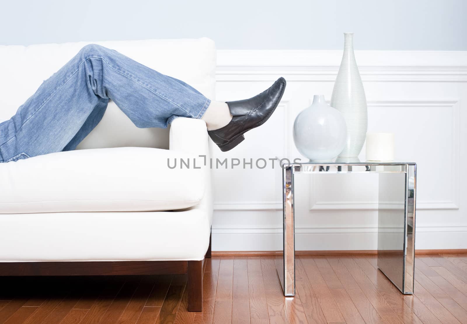 Cropped view of man reclining on white couch next to an end table holding vases, with only his legs visible. Horizontal format.