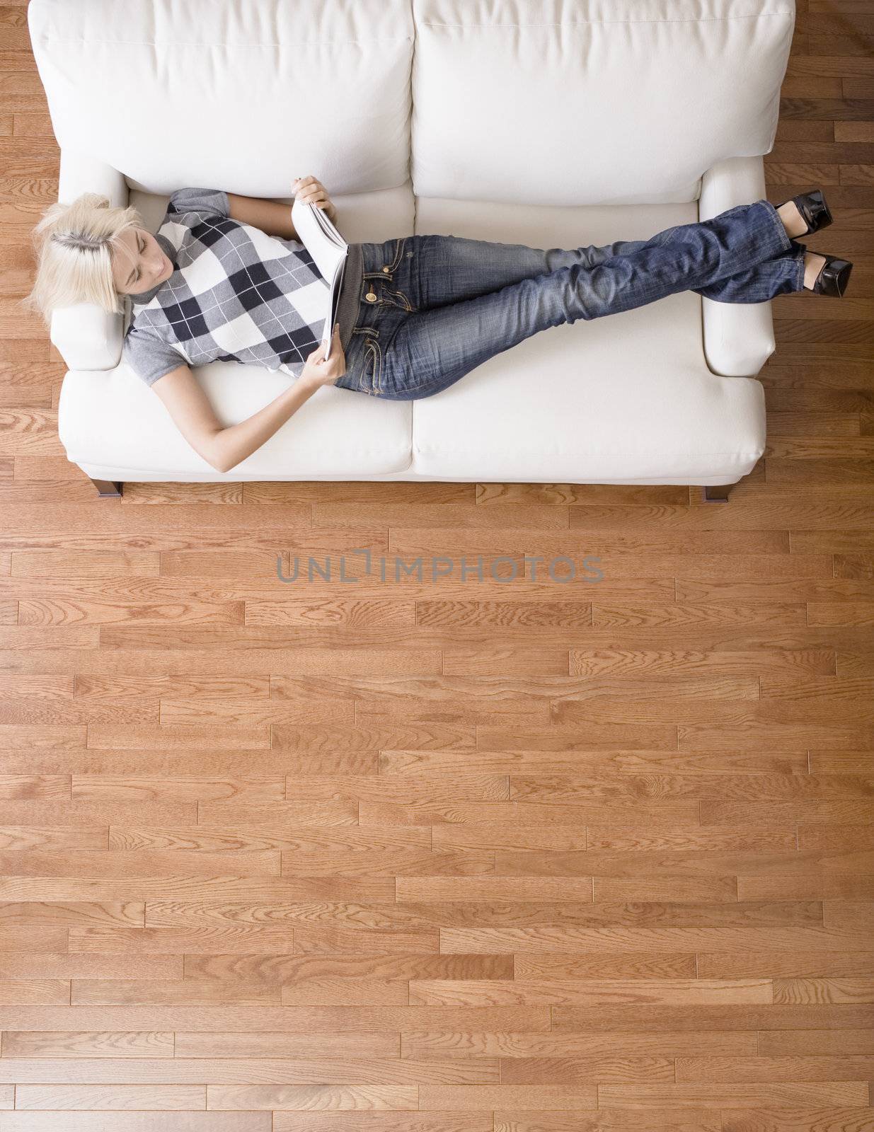 Full length overhead view of woman reclining and reading on white couch. Vertical format.