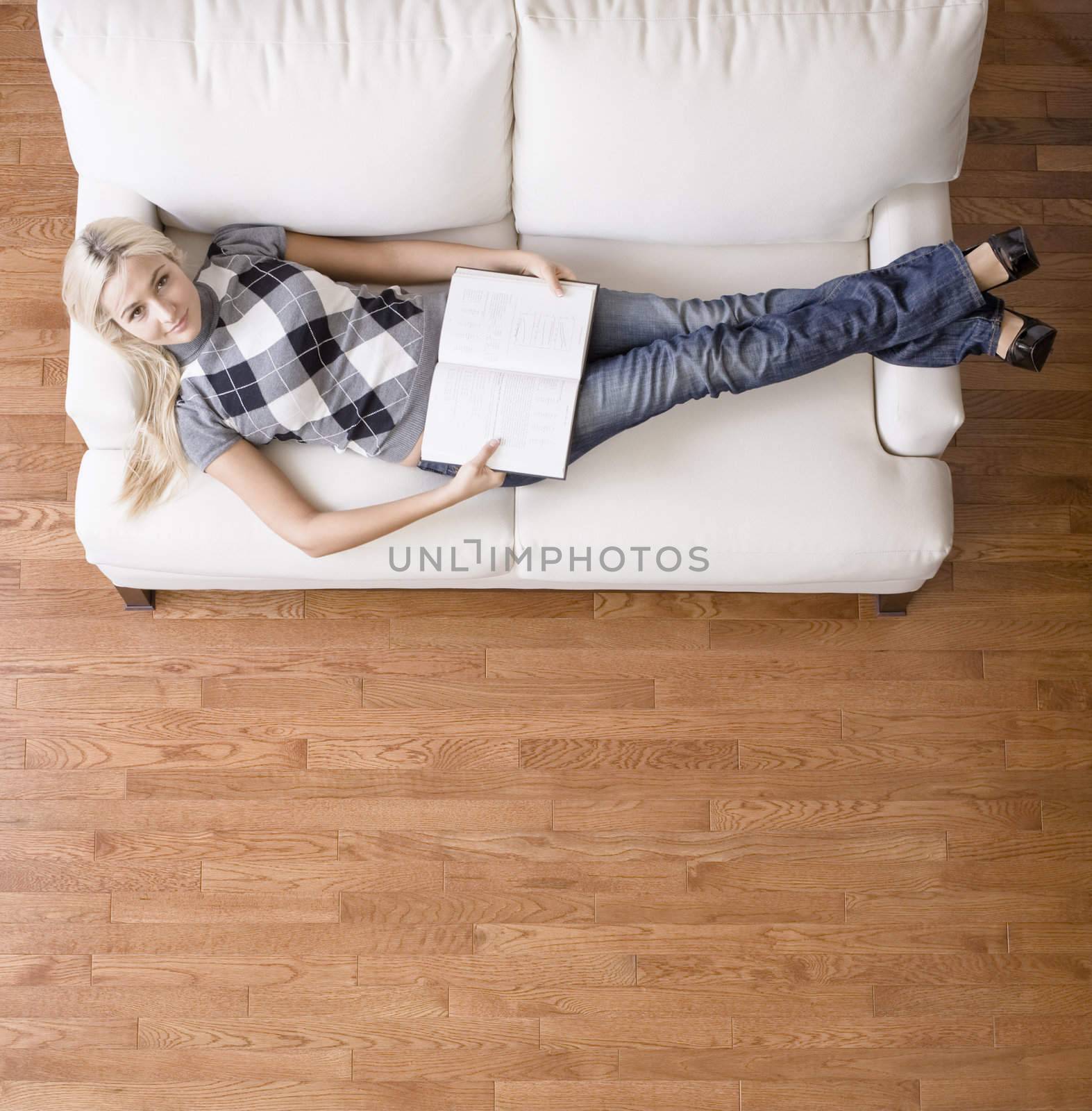 Full length overhead view of woman reclining on white couch with a book, as she looks up at the camera. Square format.