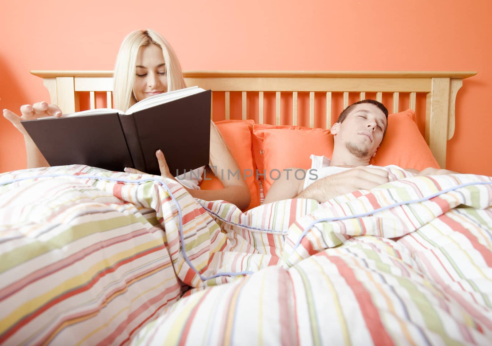 View of couple in bed, with woman reading book and man sleeping. Horizontal format.