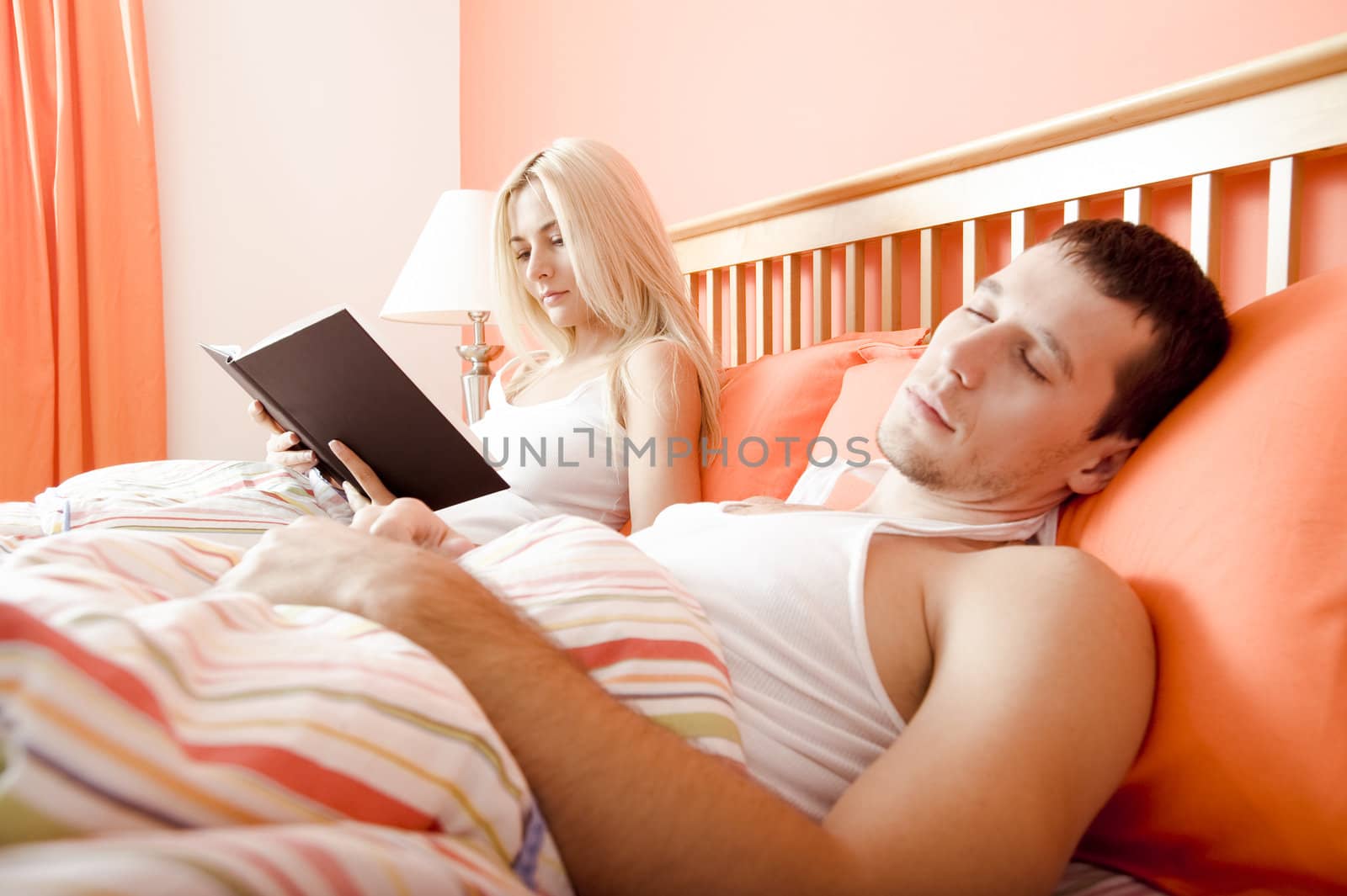 View of couple in bed, with woman reading book and man sleeping. Horizontal format.