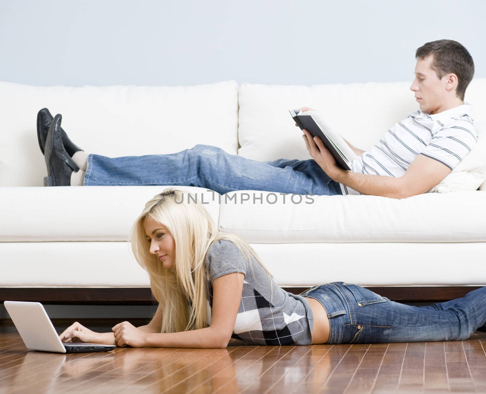 Man reads on a couch while woman stretches out on the floor with her laptop. Horizontal format.