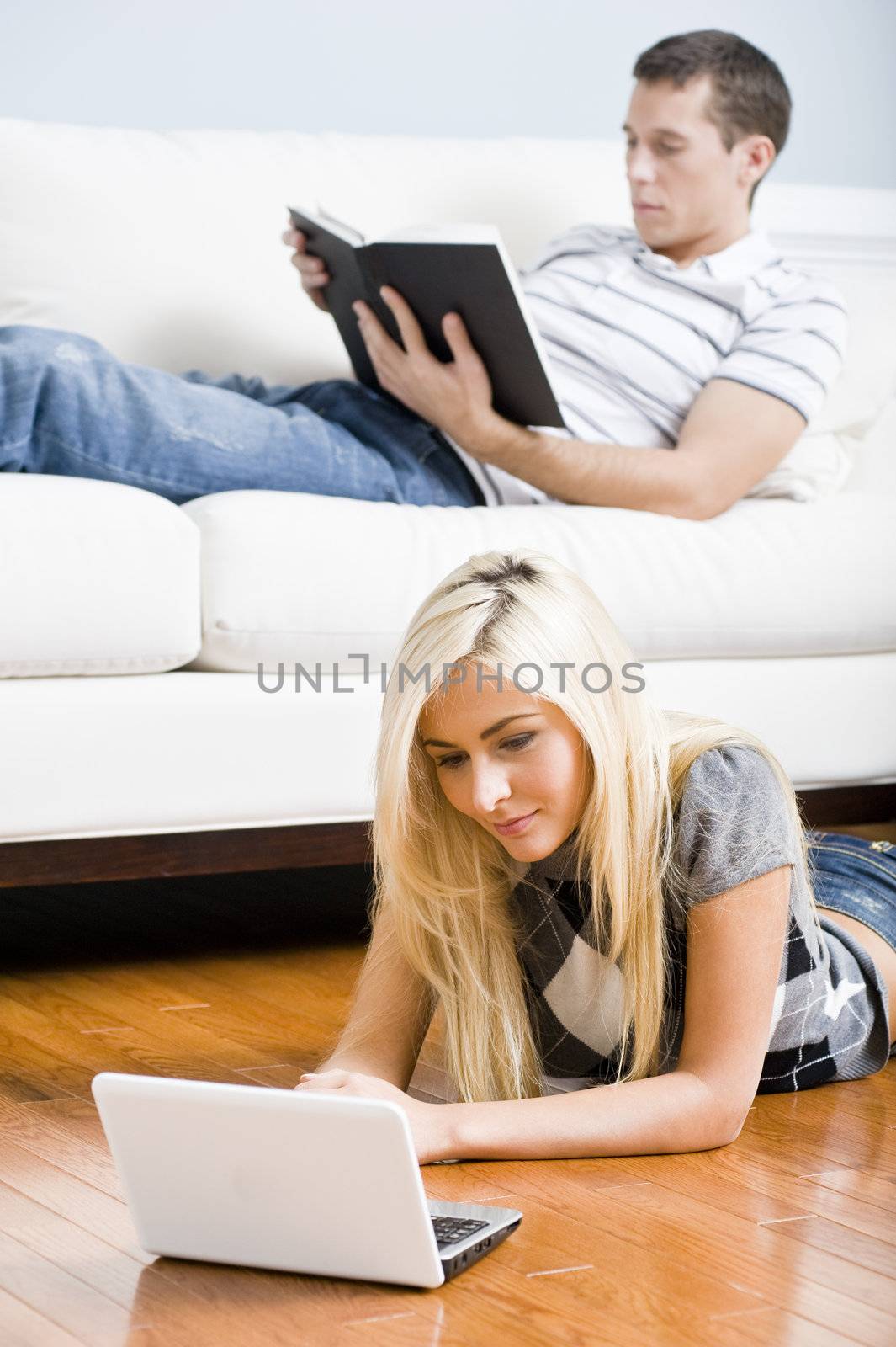 Man reads on a couch while woman stretches out on the floor with her laptop. Vertical format.