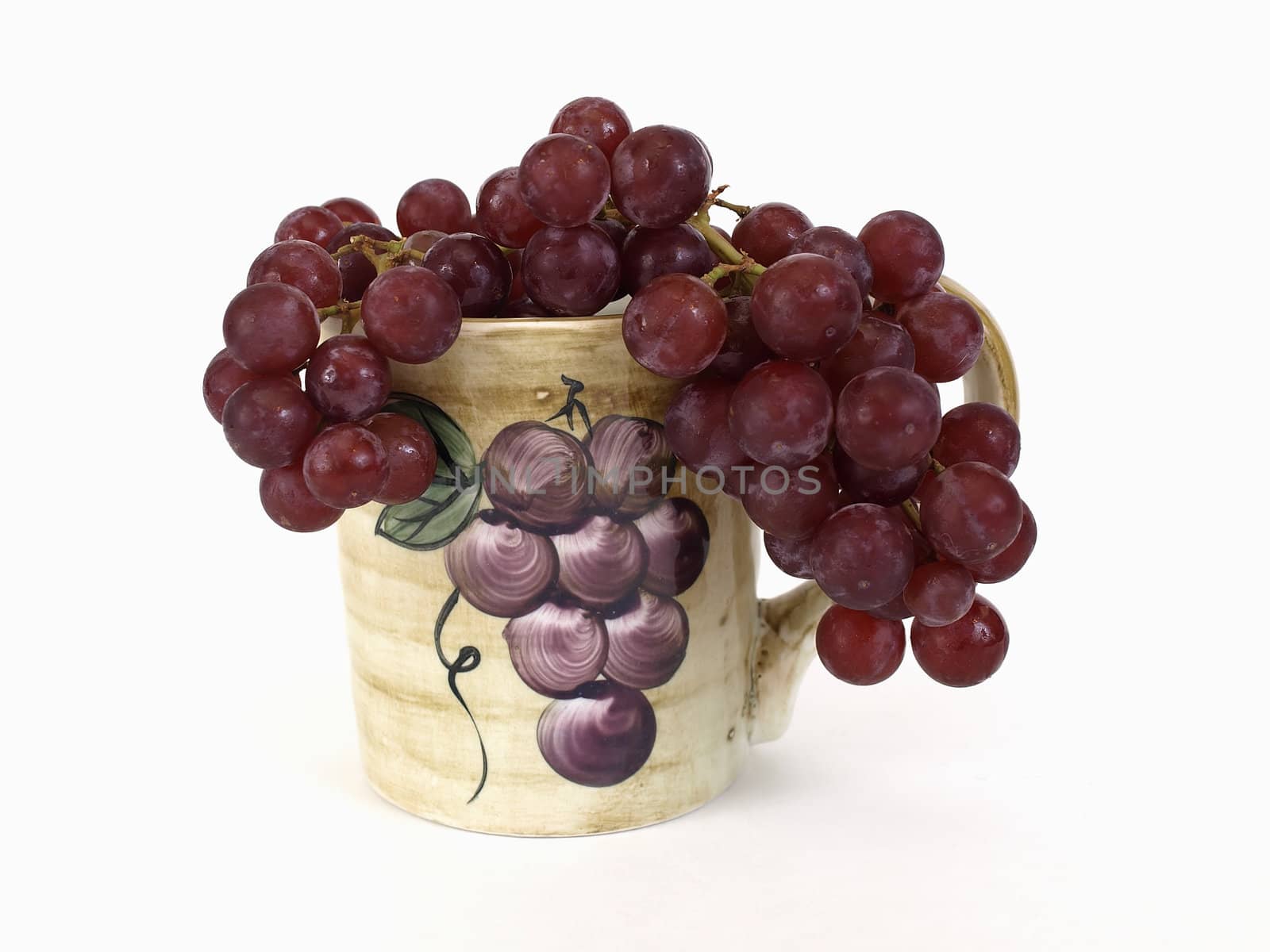 Red seedless grapes draped in a ceramic coffee cup with a grape design on it. Over white.