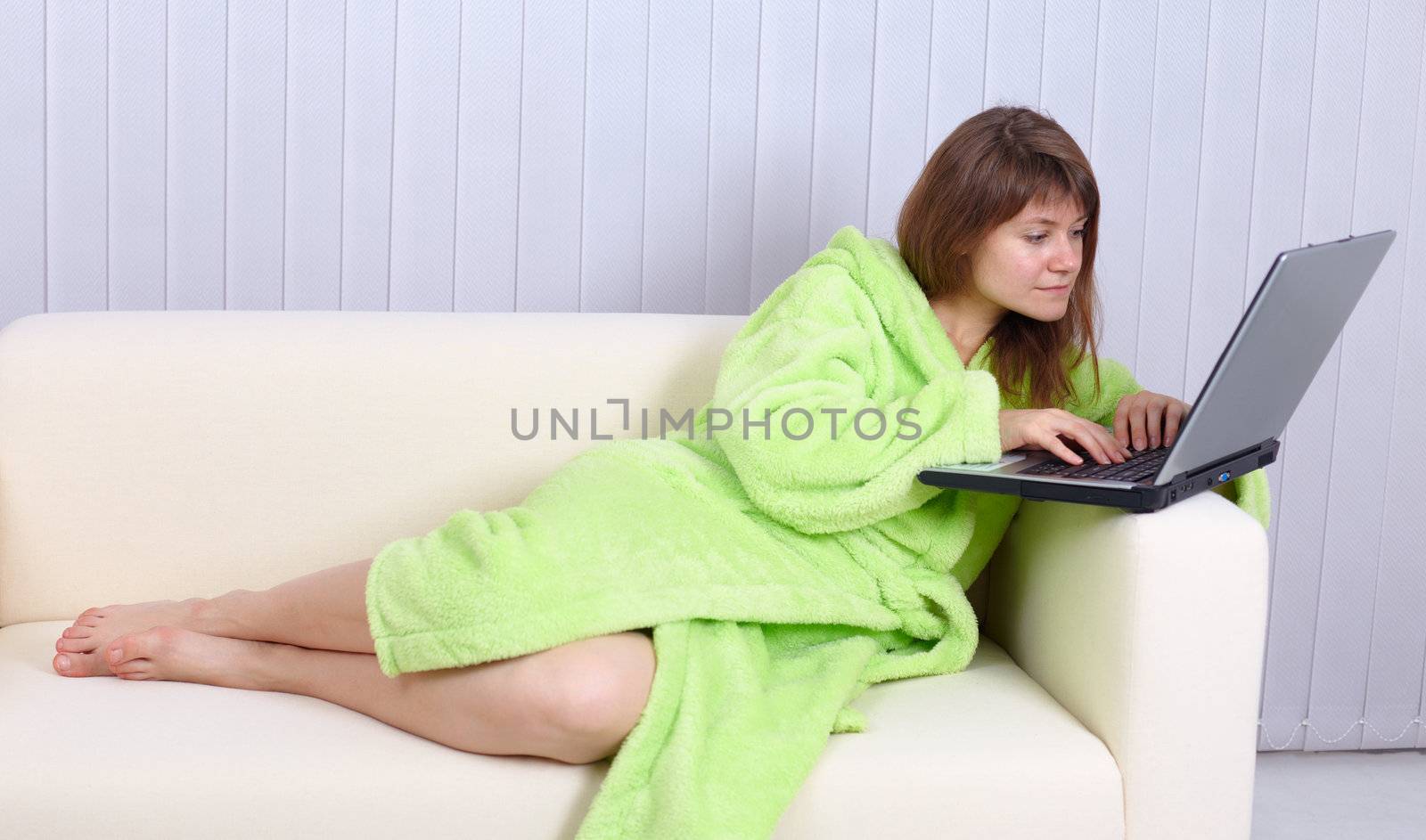 The young woman in a dressing gown reads mail by means of the laptop lying on a sofa