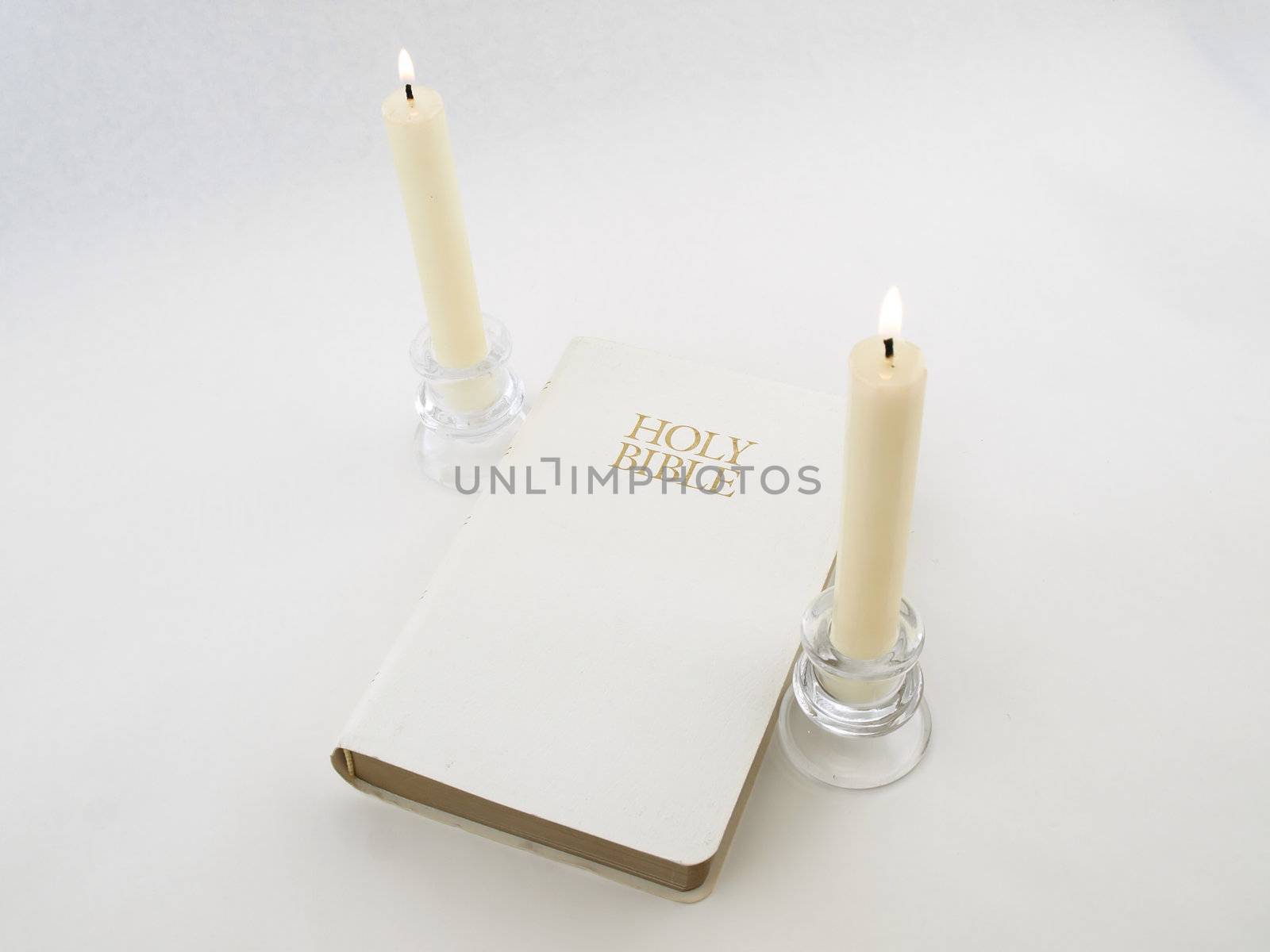 A white Holy Bible between two white lit candlesticks on a white background.
