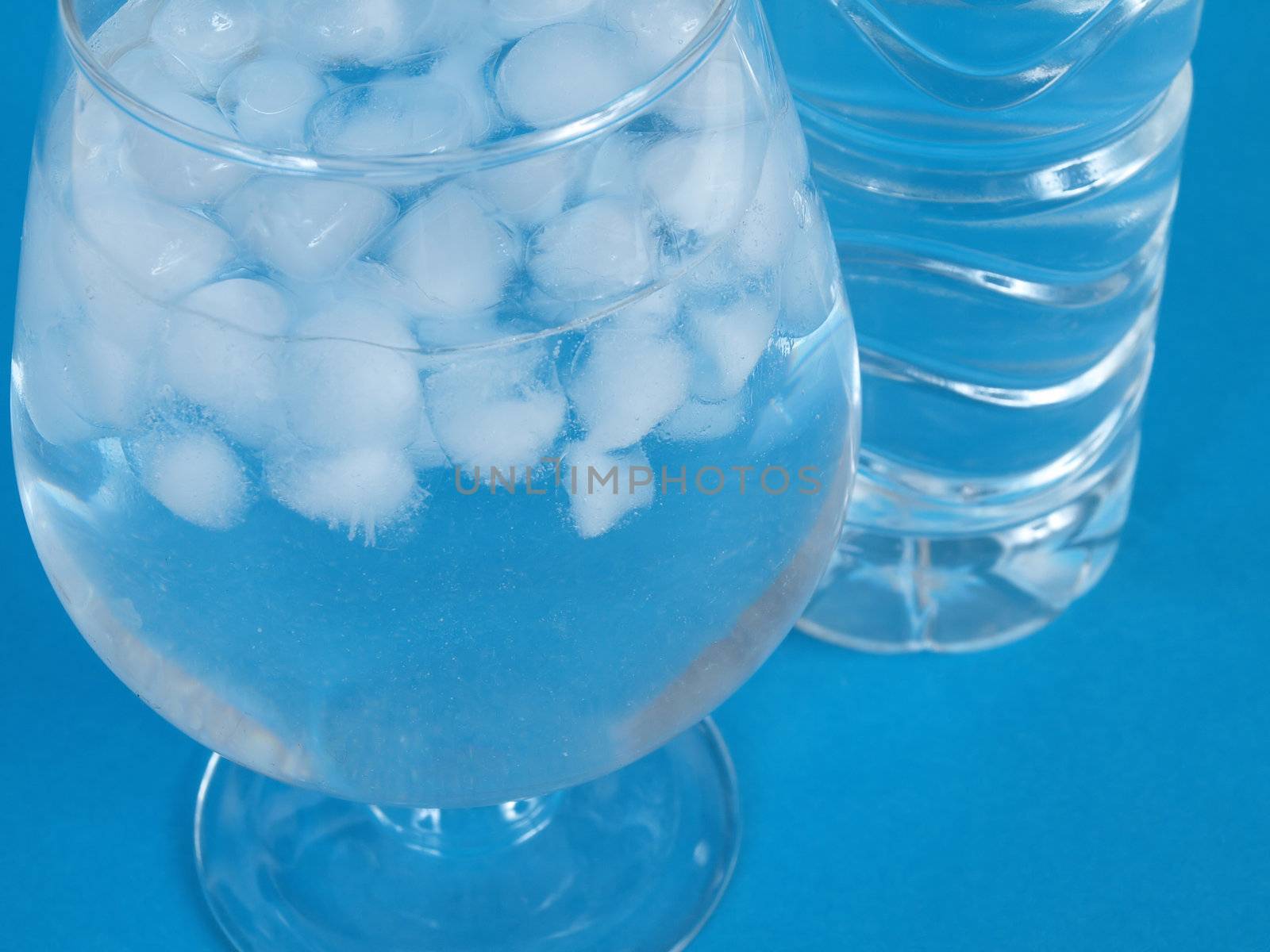 An ice filled glass with water next to a plastic water bottle, over a blue background.