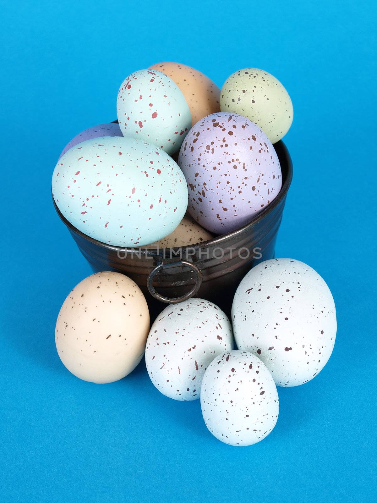 Flecked pastel colored eggs fill a metal pail, a few eggs laying to the side, over a blue background.