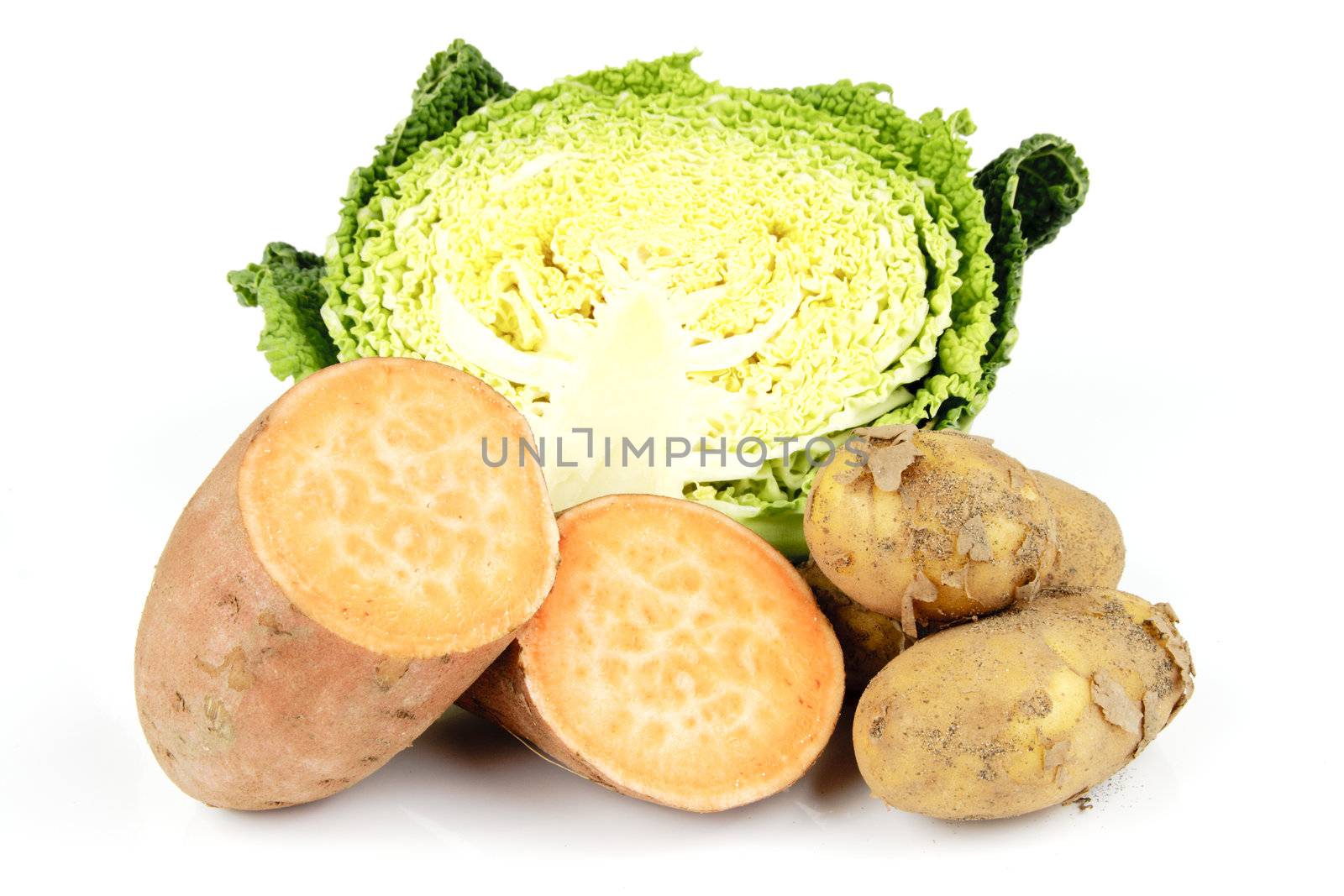 Half a raw green cabbage with a single sweet potato cut in half and a pile of brown potatoes on a reflective white background