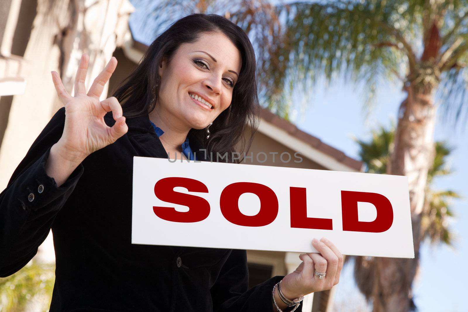 Happy Attractive Hispanic Woman Holding Sold Sign In Front of House.