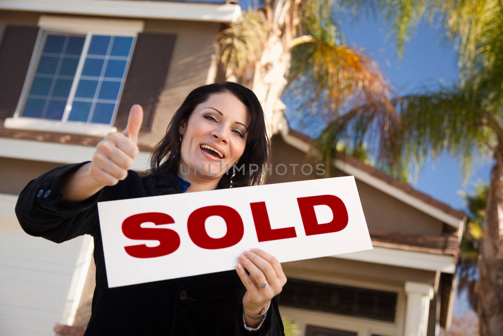 Happy Attractive Hispanic Woman with Thumbs Up Holding Sold Sign In Front of House.