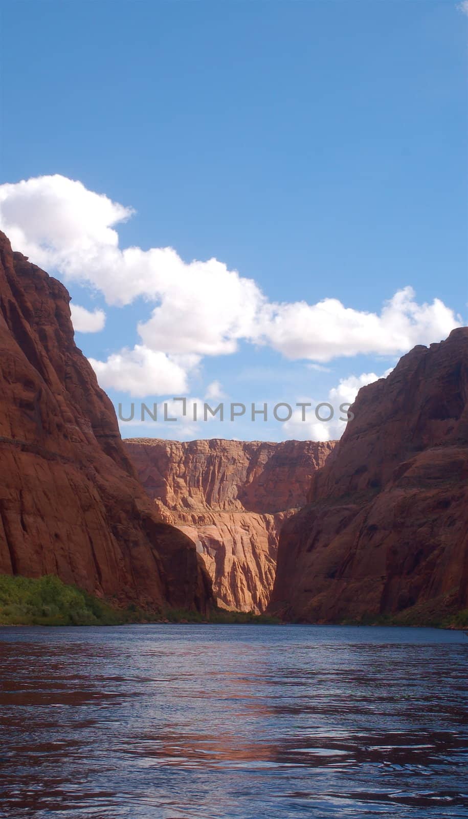 View from the Colorado River, looking up at the red rock walls of the Grand Canyon into a blue cloudy sky.