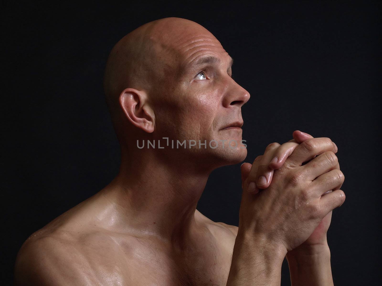 A bald shirtless man clasps his hands in prayer, eyes pleading skyward, over a black background.