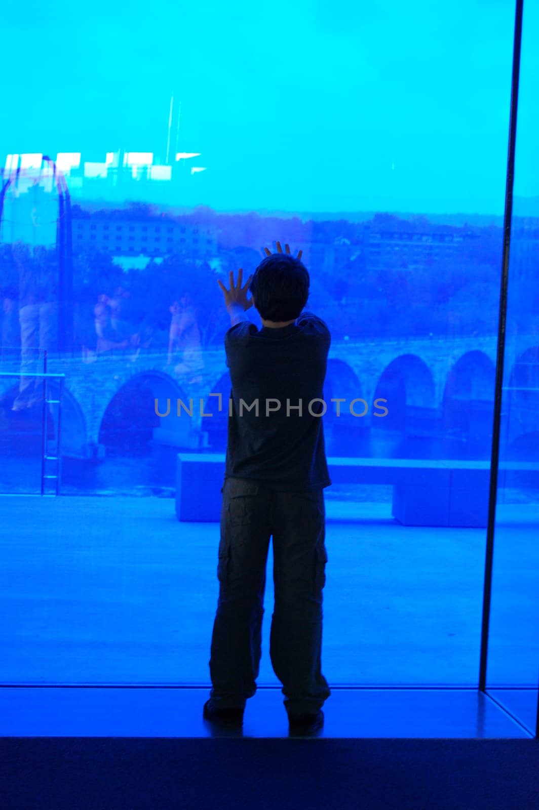 Boy looking through blue glass window onto city below.  by RefocusPhoto