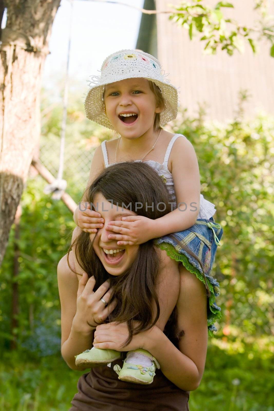 Little laughing girl sits on shoulders at other laughing girl in the afternoon in a garden
