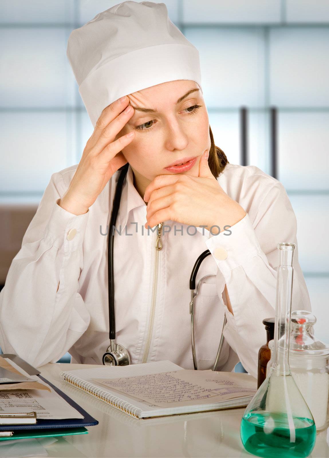 Portrait of the tired woman-therapist in a white uniform with a stethoscope, sitting near a table with records and medicines
