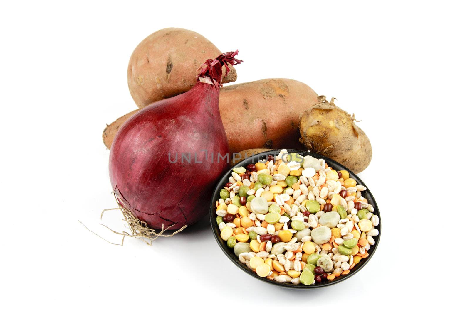 Two raw unpeeled sweet potatoes with a red onion and a black dish of soup pulses on a reflective white background