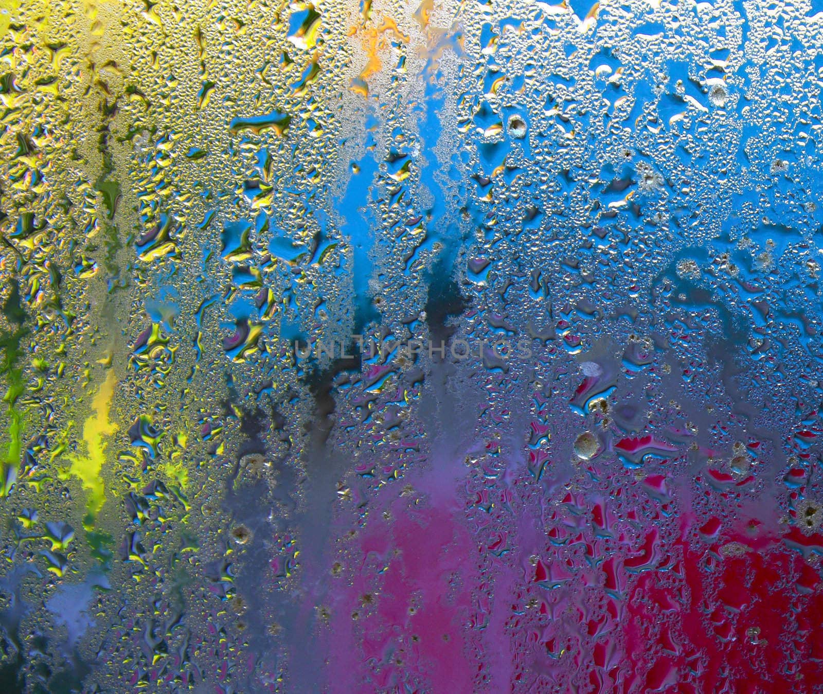 Condensation on window glass with blurred colors, primarily yellow, blue and red, in the background.