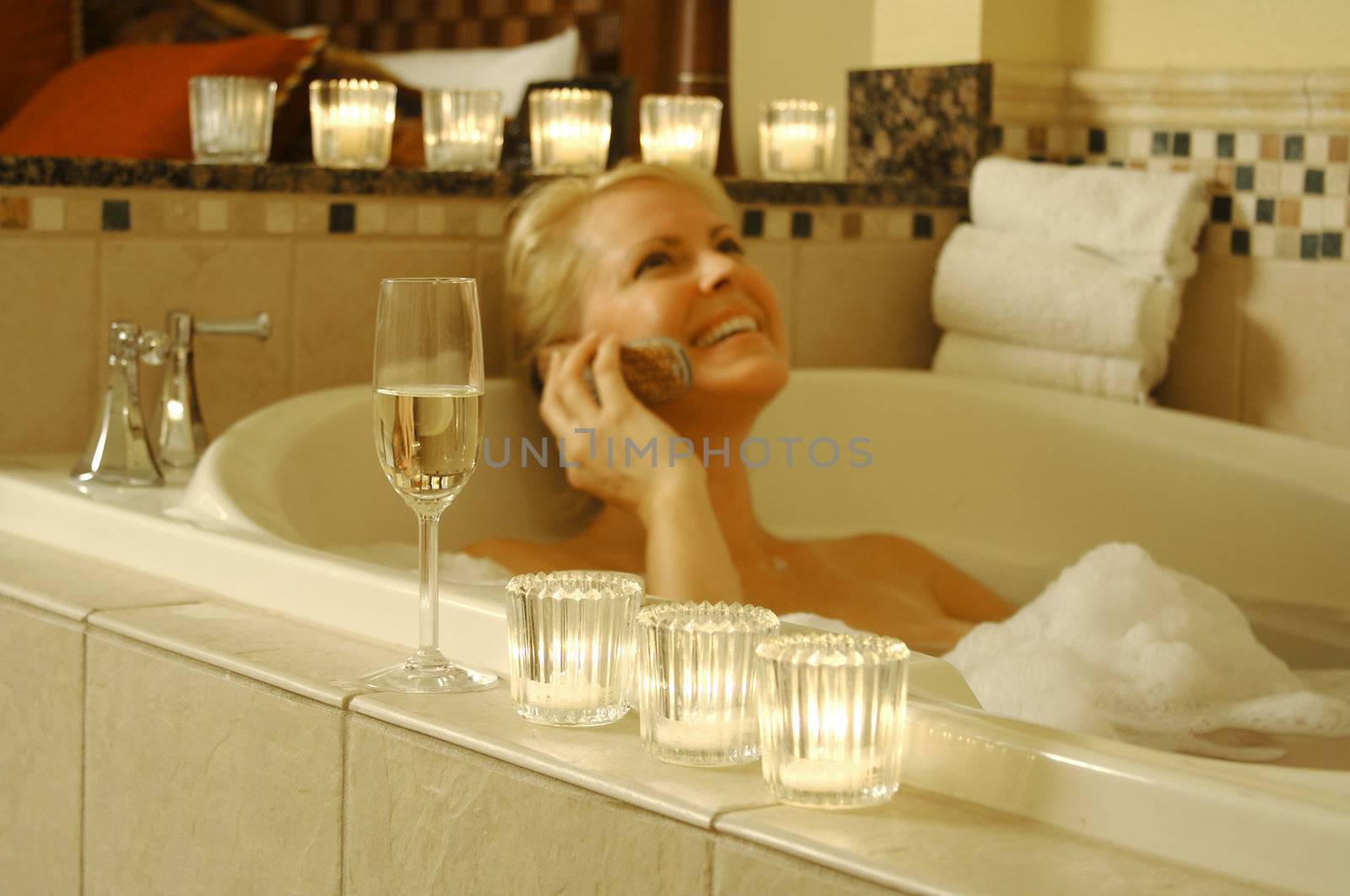 Beautiful Woman using cell phone in bubble bath with sparkling wine and candles surrounding her.