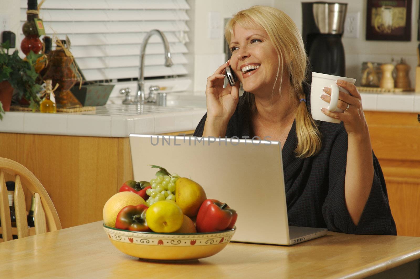 Woman smiling, in her kitchen on cell phone sitting in front of laptop.