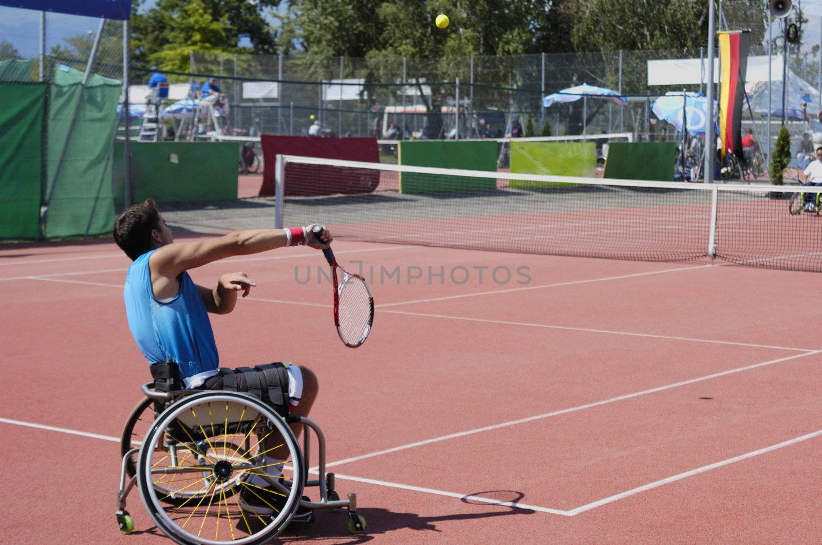 A wheelchair tennis player during a tennis championship match, serving. The ball, with motion blur, is visible centre top.