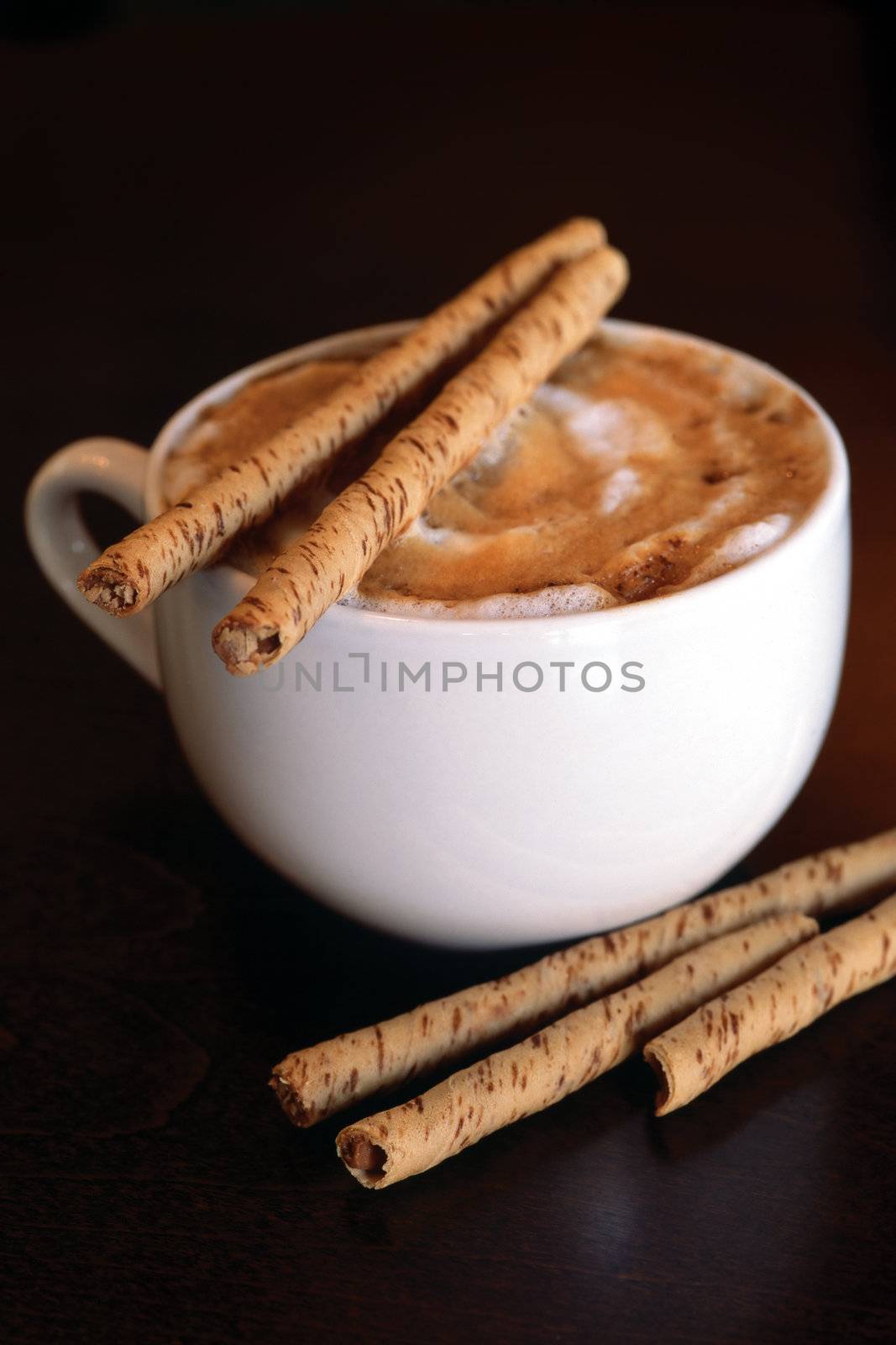 Cinnamon sprinkled hot cappuccino with chocolate sticks on a dark wood table.
