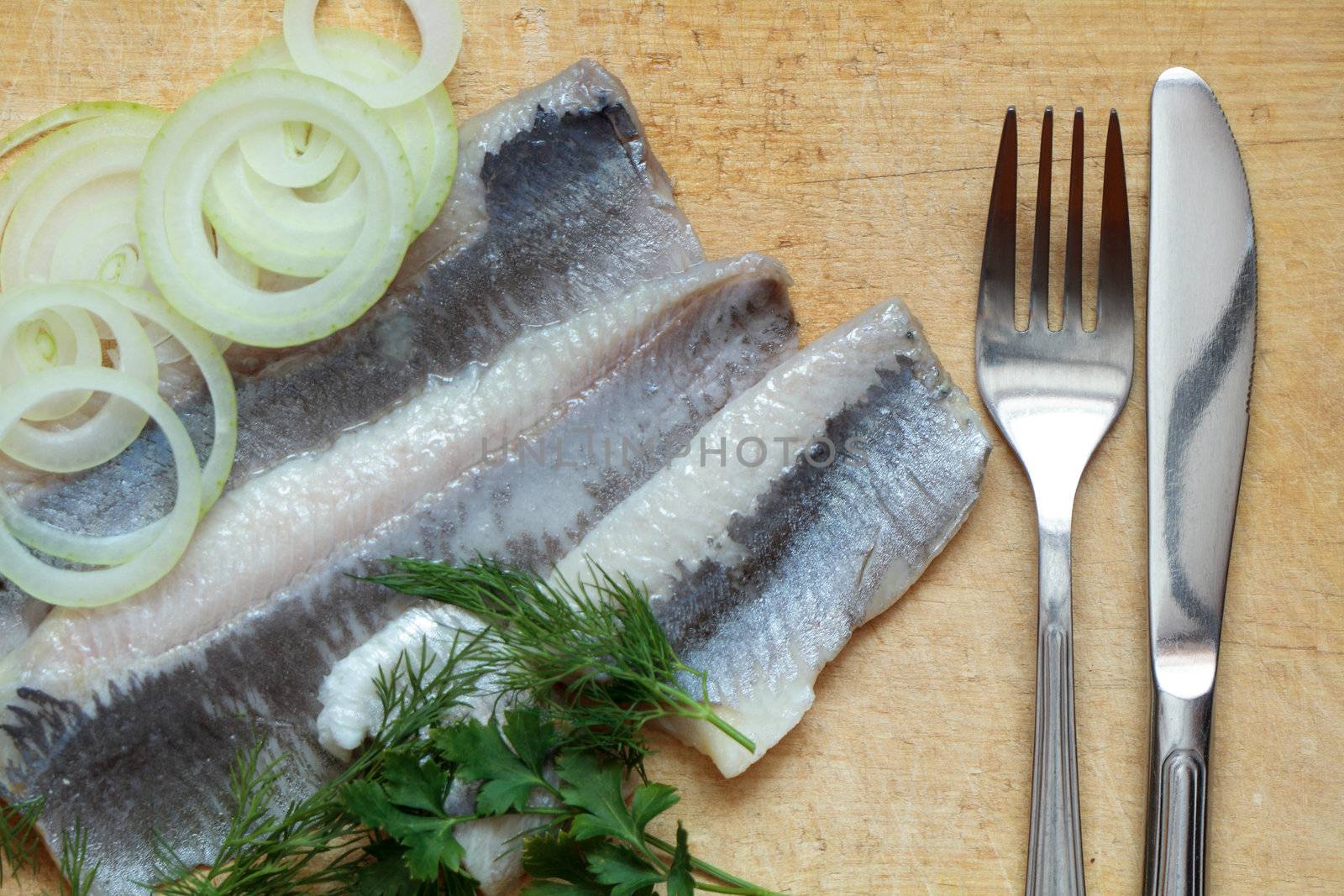 Still life with sliced herring, onion, greens, fork and knife on wooden background