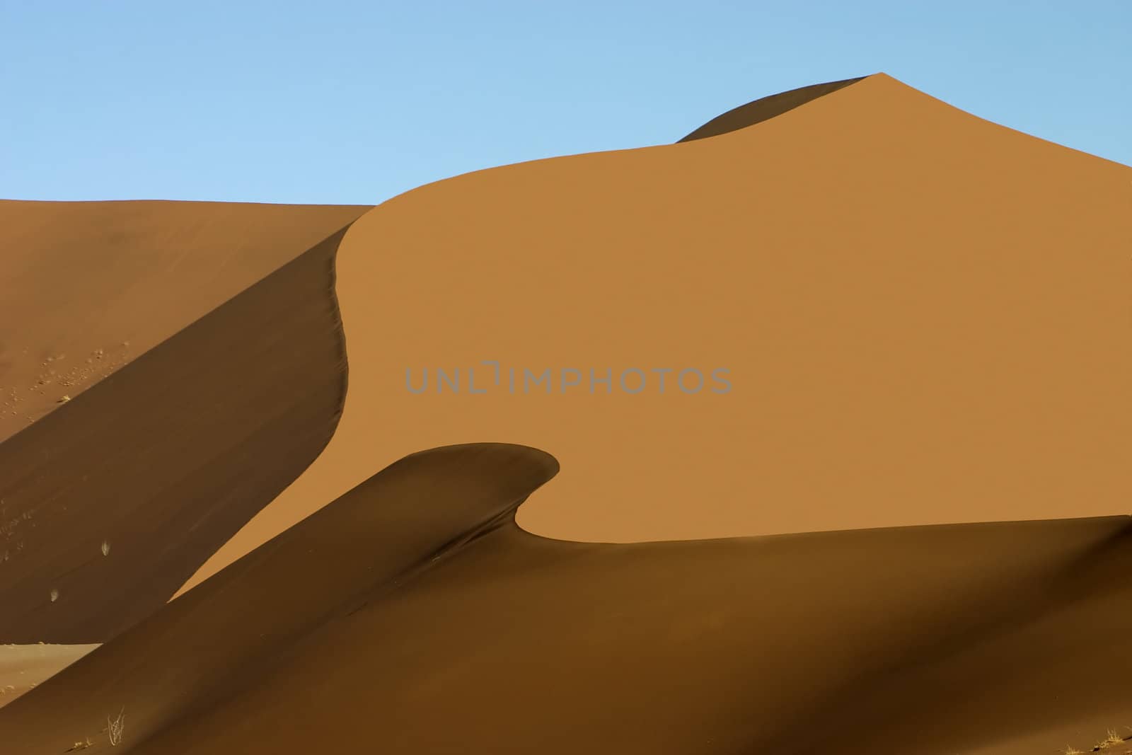 A large orange sand dune with wave like shadows to the bottom and side against a light blue sky