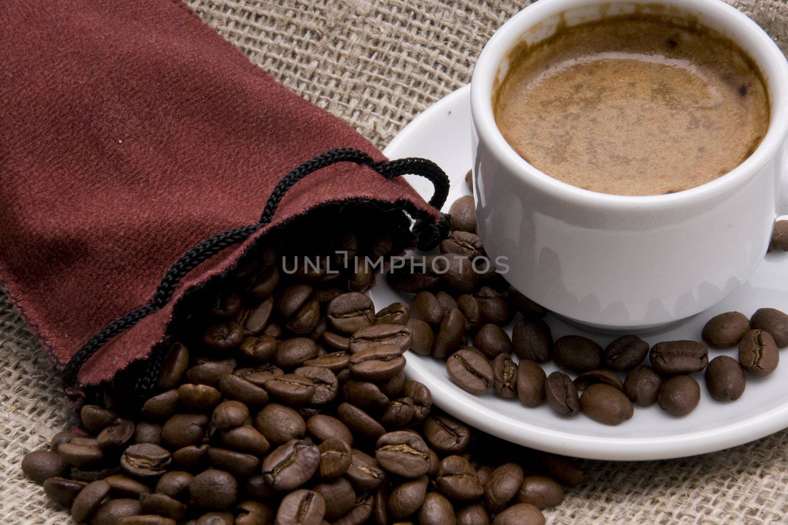 A bag filled with coffe-beans and white cup of coffee with saucer is standing on jute-fabric.
