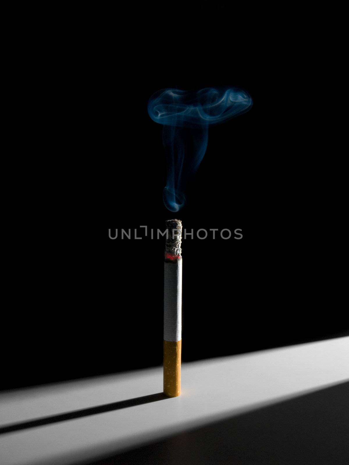 A single smoking cigarette standing in a narrow corridor of light. A conceptual image about the smoking habit.