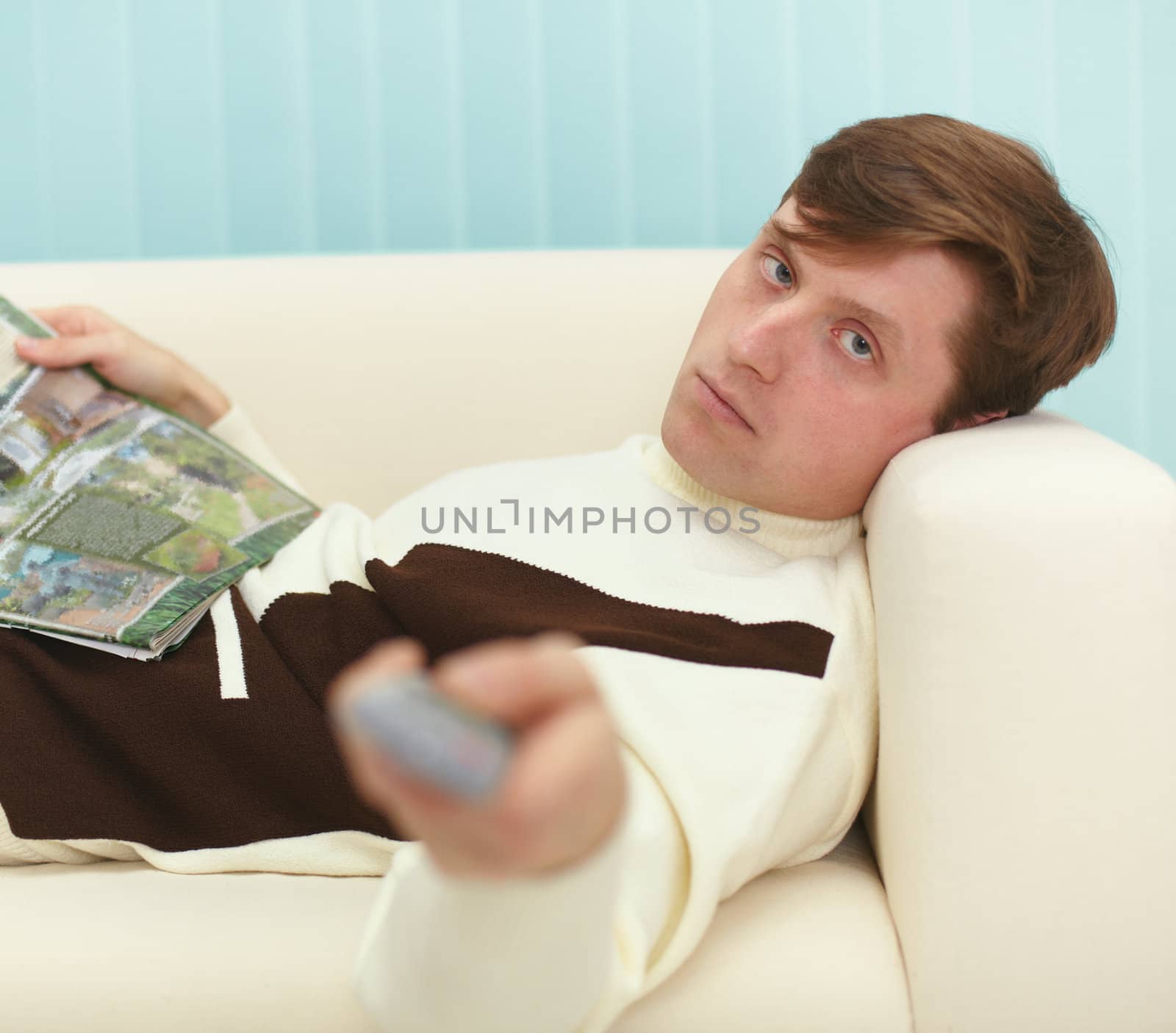 A young man, lying on the couch with a magazine and the TV remote control