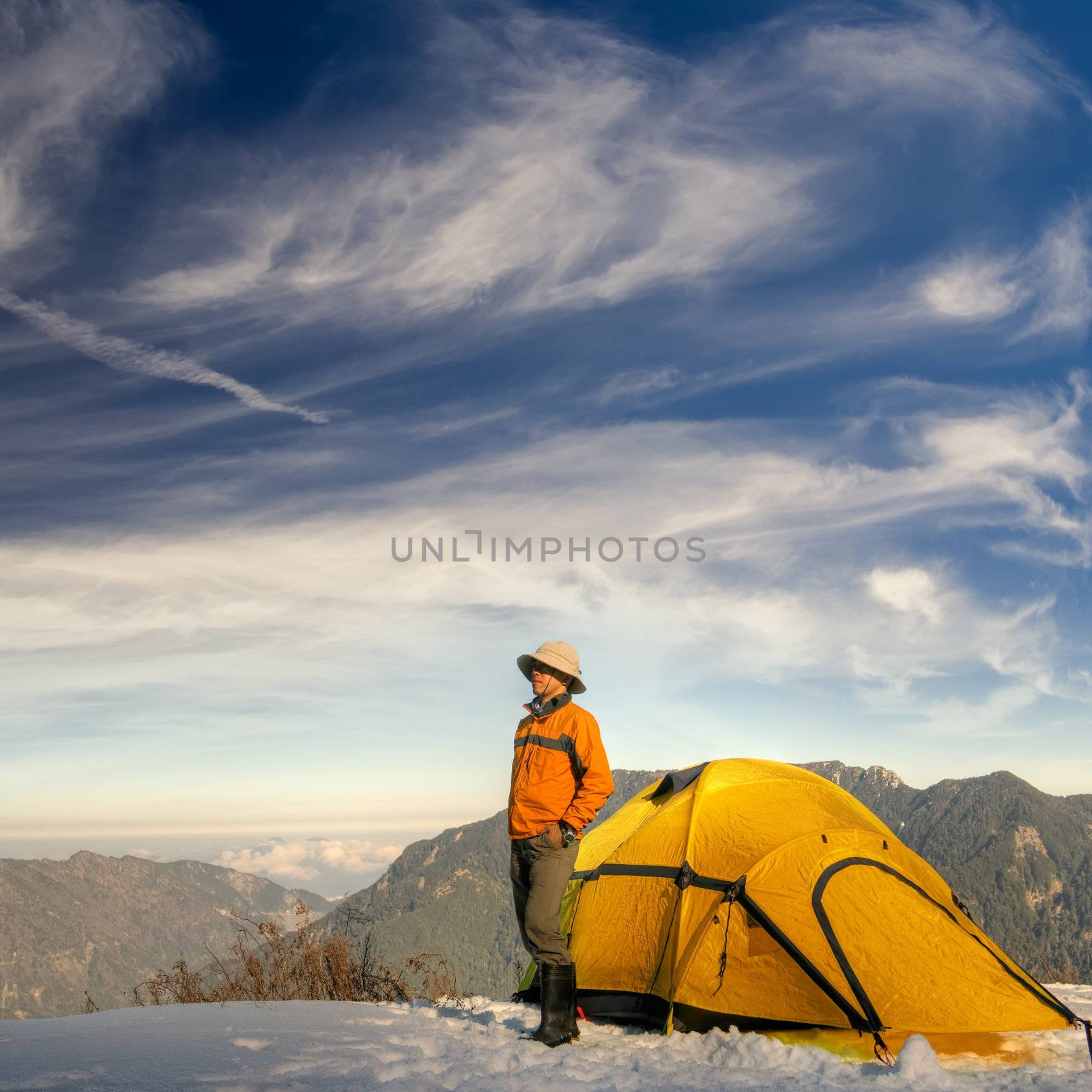 Man stand with yellow tent on snow hill against dramatic sky.