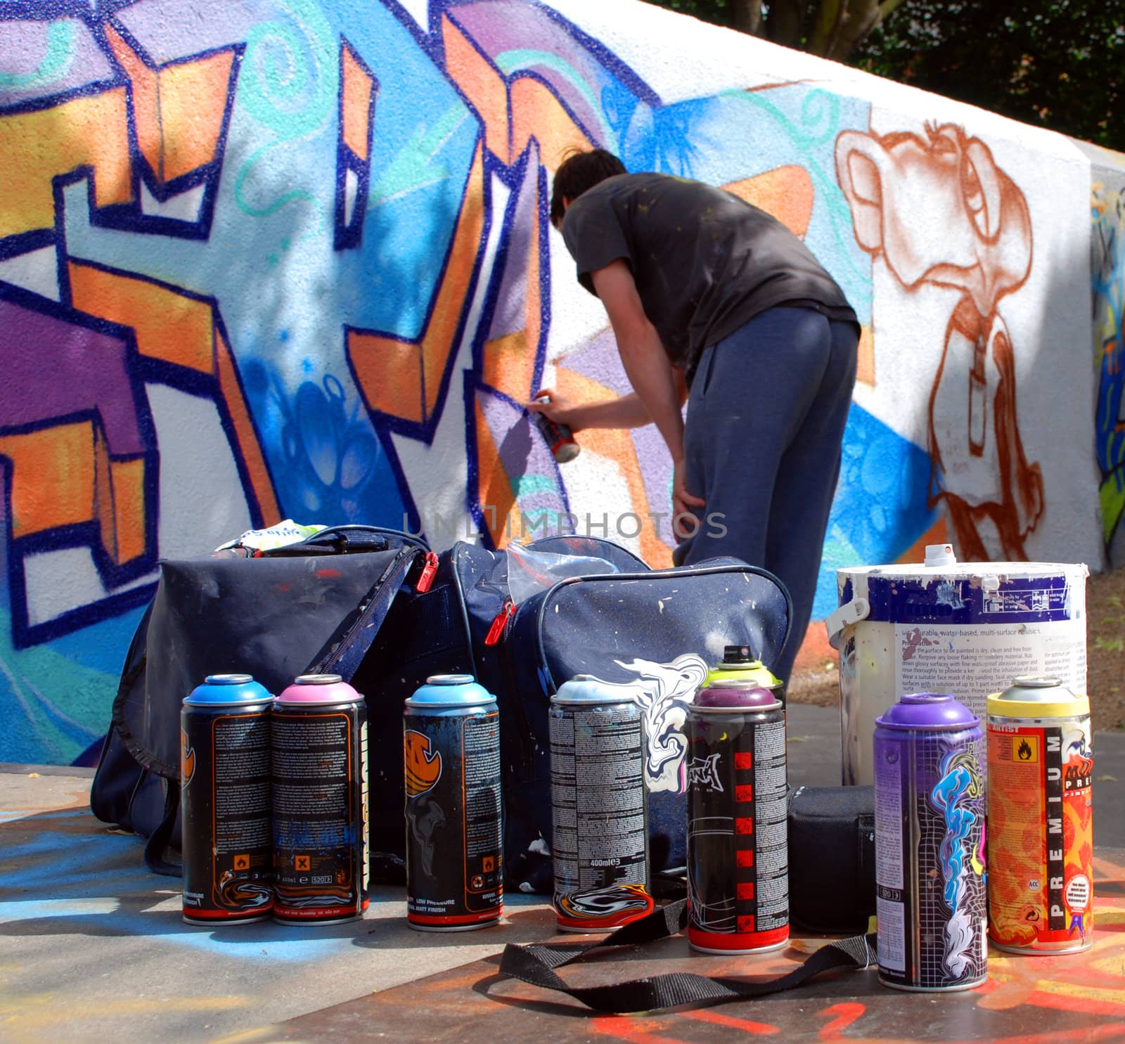 A photograph of an unidentifiable graffiti artist spray painting a public wall, with paint and cans in the foreground