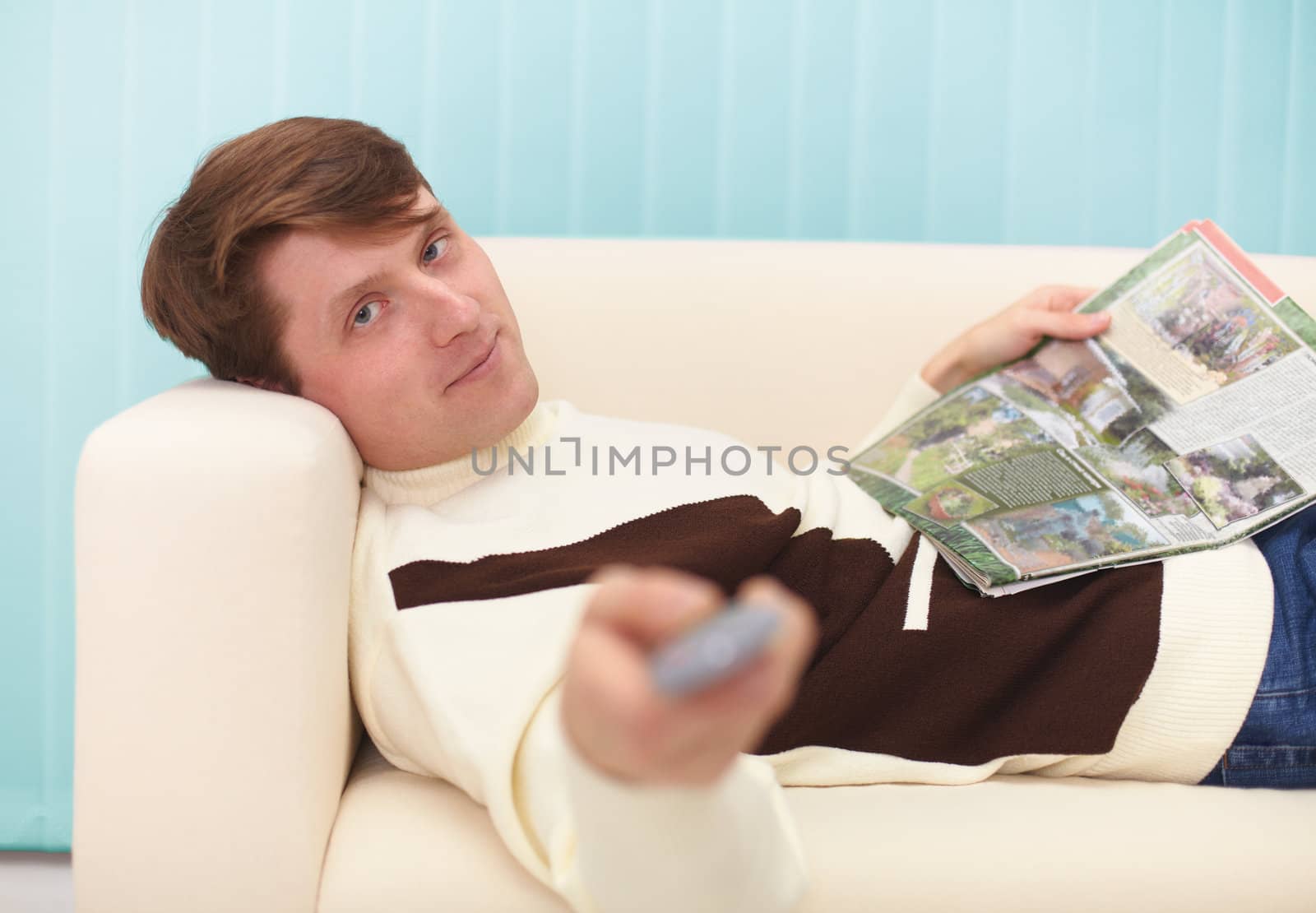 A smiling man, lying on the couch with a magazine and the TV remote control