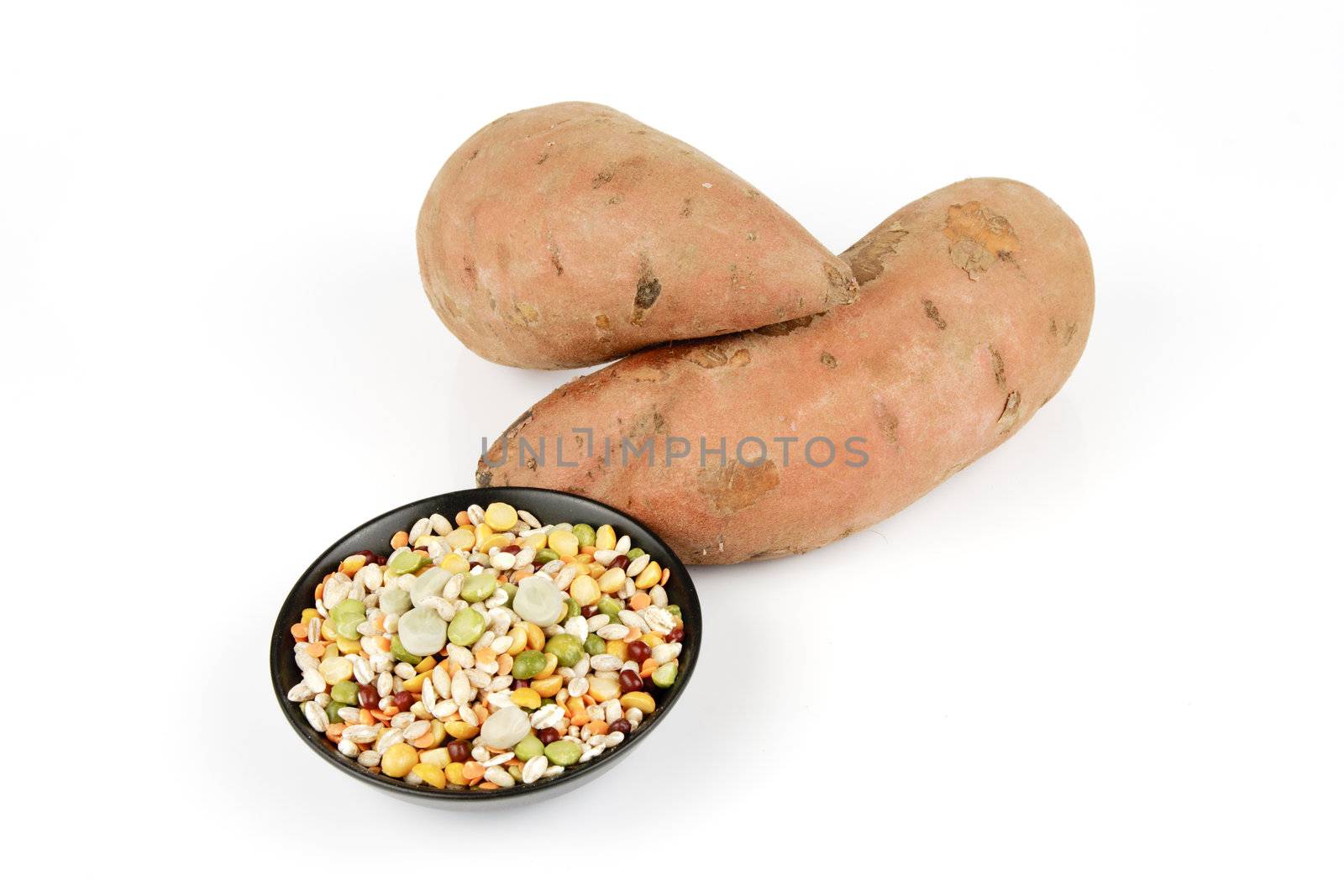 Two raw unpeeled sweet potatoes with a small black dish of soup pulses on a reflective white background