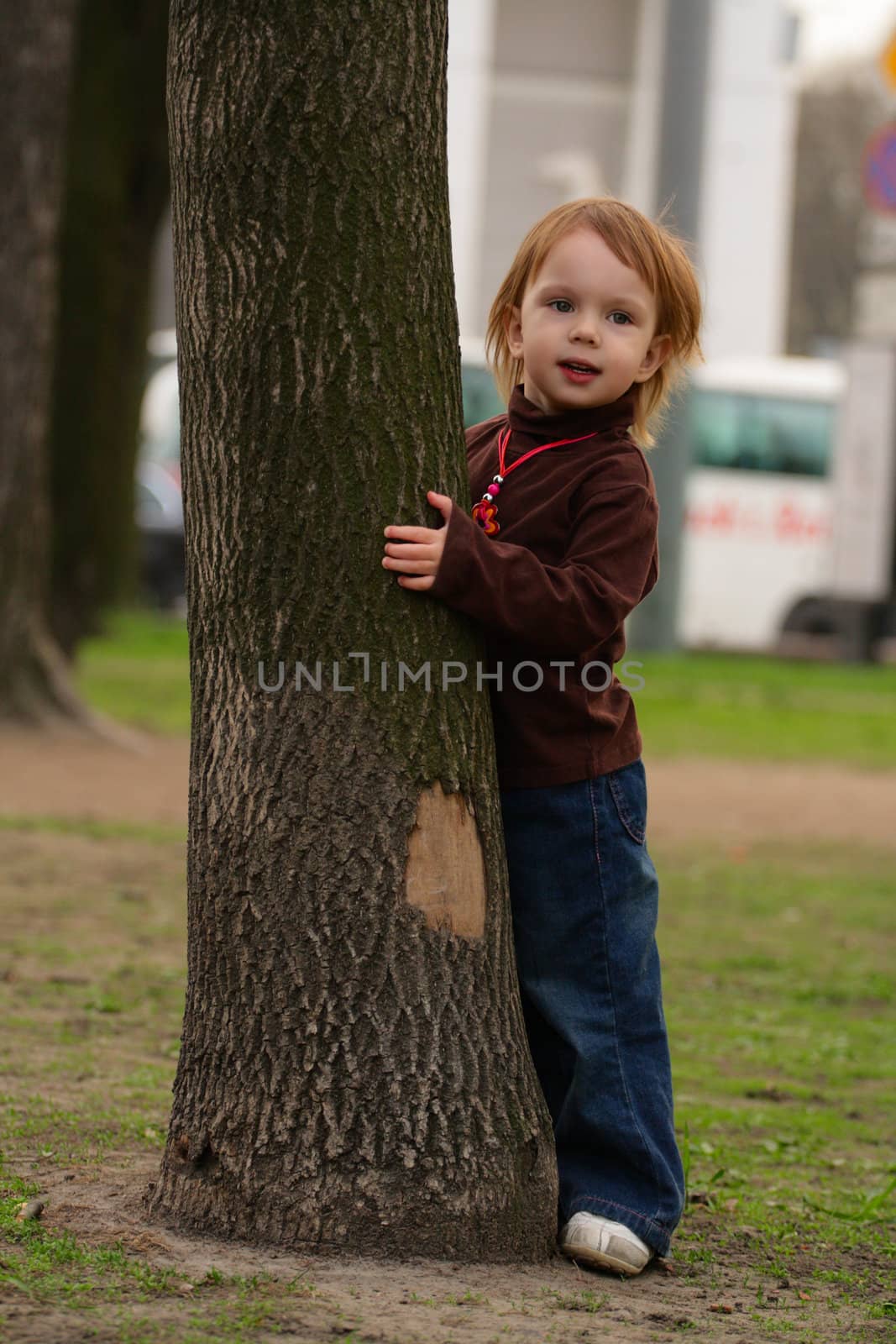 Little beauty girl playing hide-and-seek at the park. She hideing round the big old tree with split off part of bark.