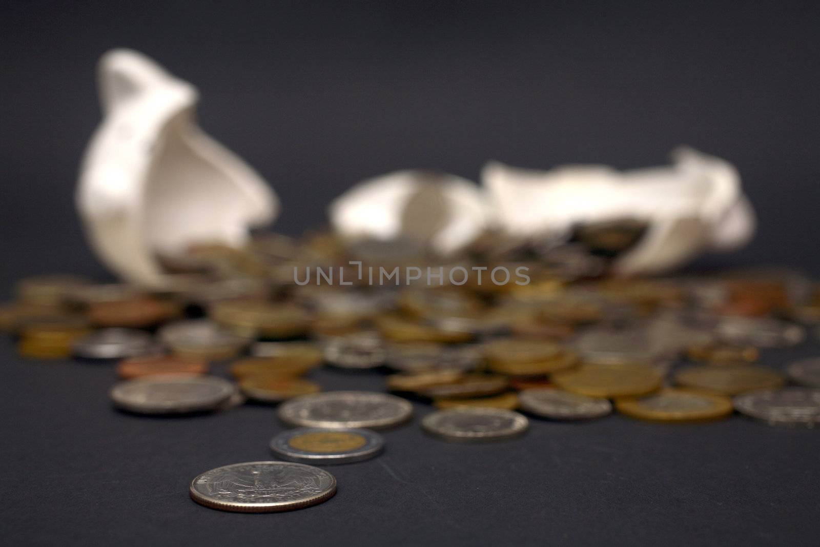 A broken piggy bank isolated on a dark background with loads of coins from around the world.